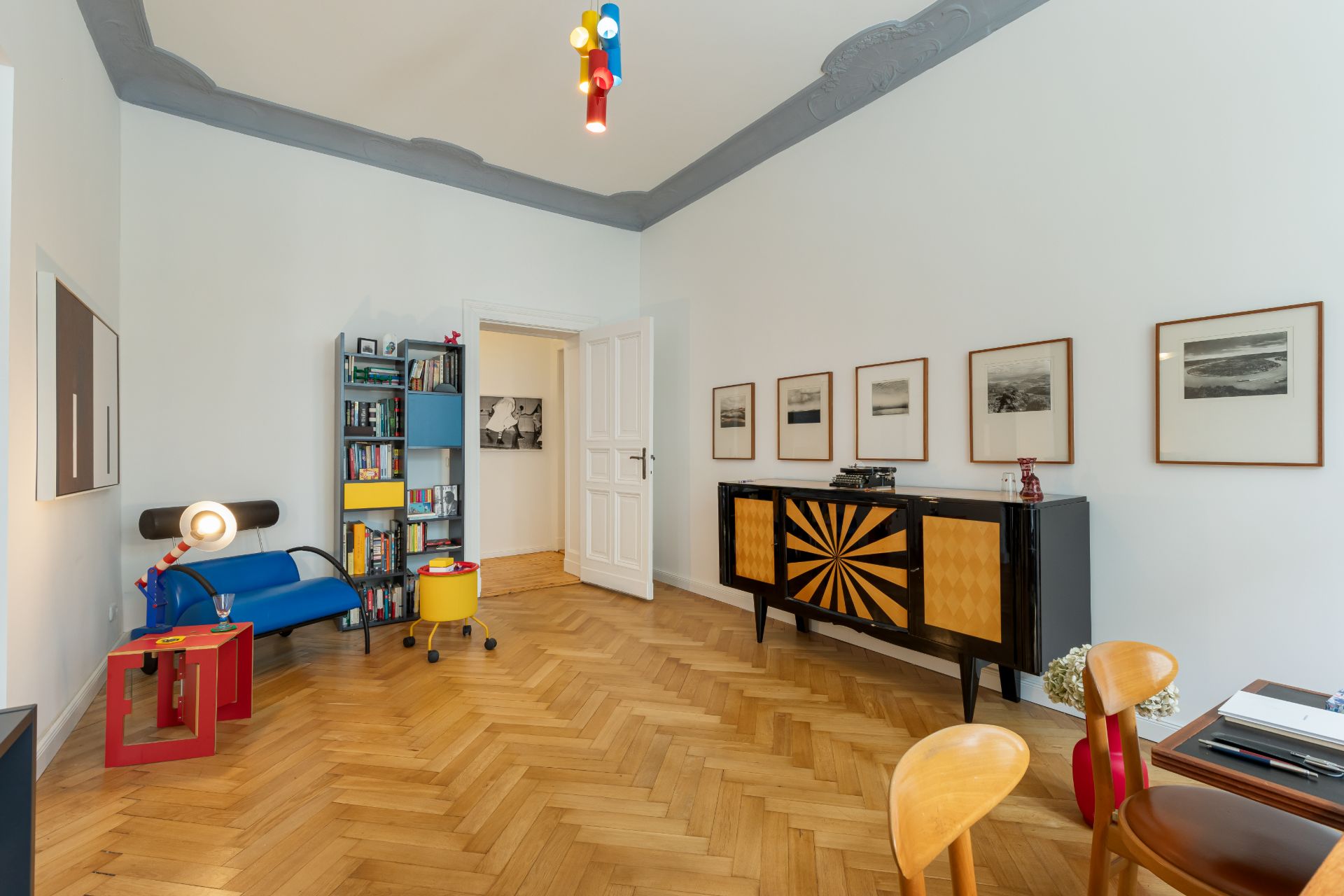 Wohnzimmer mit Parkettboden, bunter Lampe, Regal, Sofa und Kommode mit geometrischem Muster.