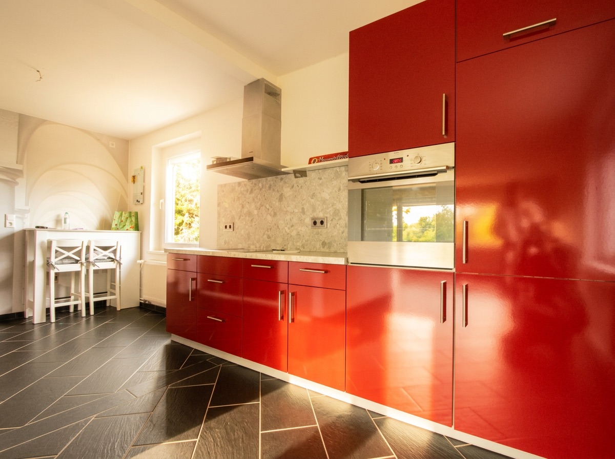 Moderne Küche mit roten Hochglanzschränken, Backofen und Esstisch mit zwei weißen Stühlen.
