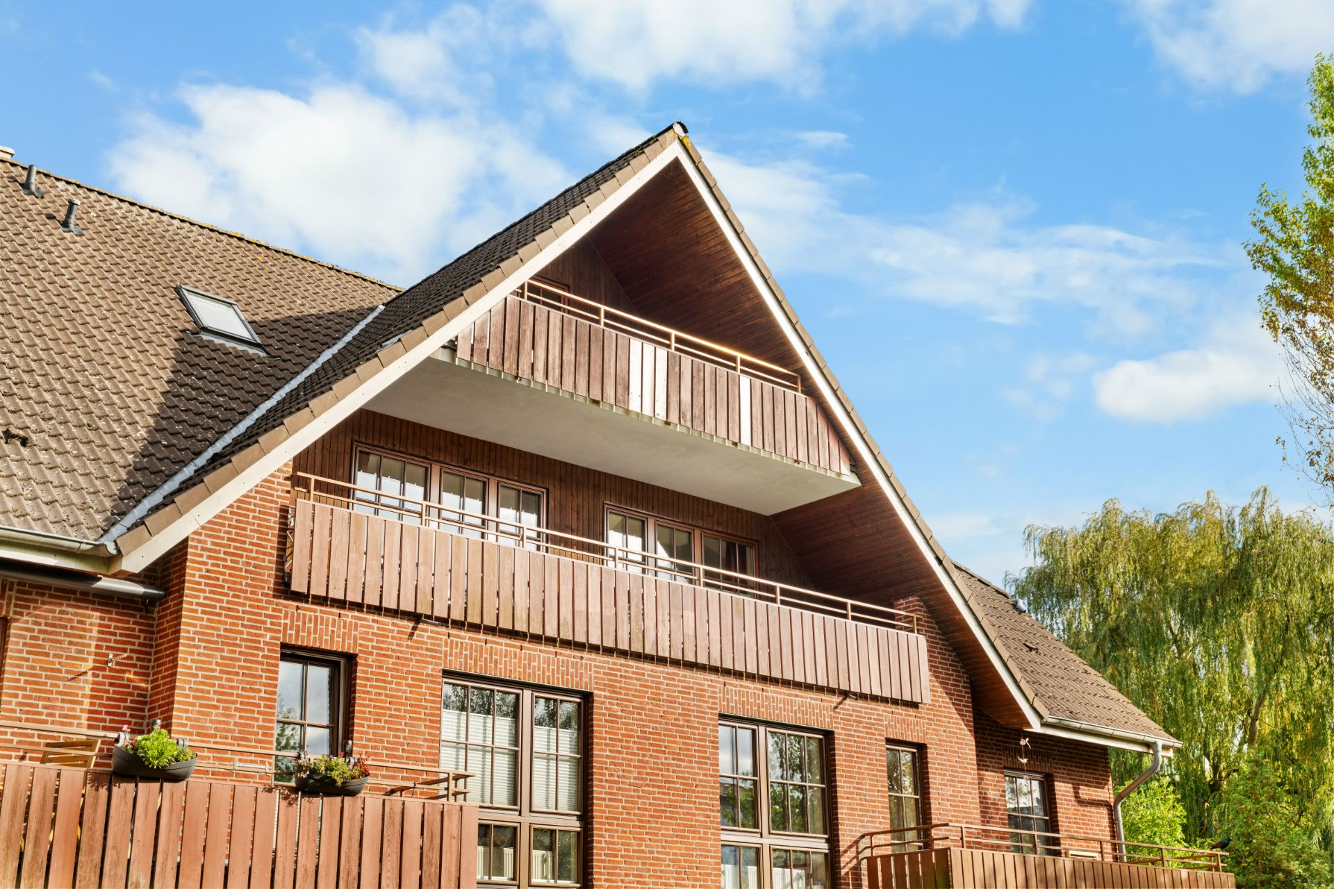 Backsteinwohnhaus mit großem Balkon, Holzgeländern und Schrägdach unter blauem Himmel.