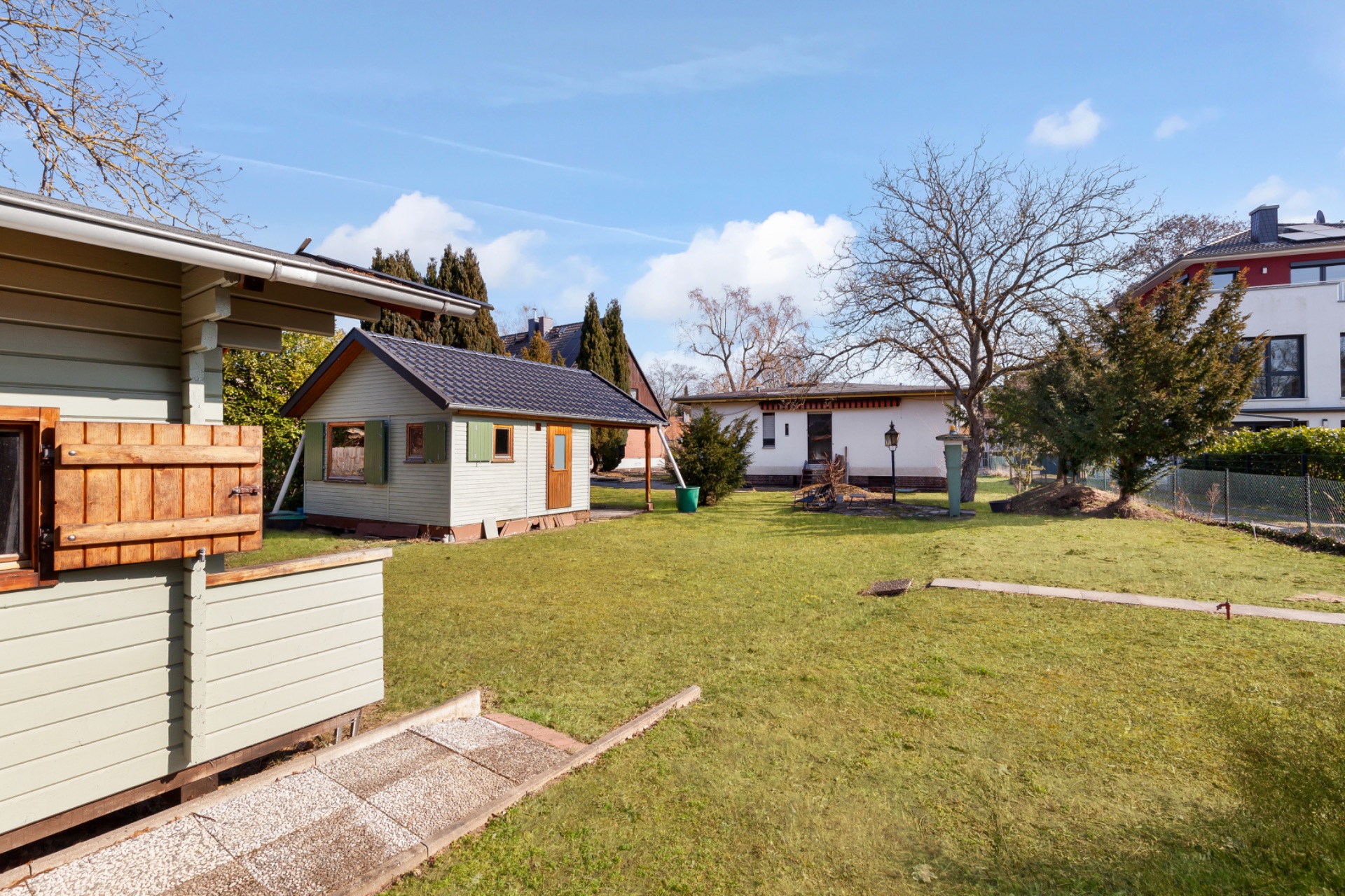 Garten mit zwei kleinen Holzhäusern und Rasenfläche, umgeben von Bäumen und anderen Gebäuden.