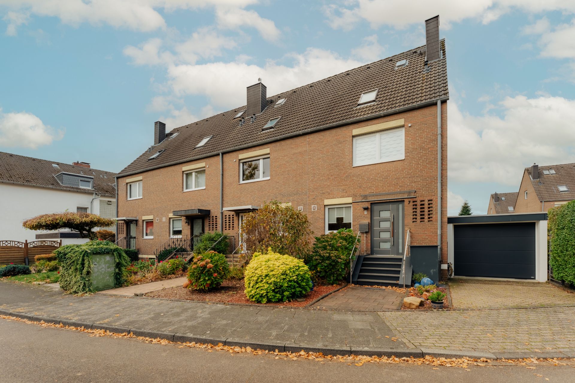 Reihenhaus aus Backstein mit Vorgarten, Garagenauffahrt und bepflanzter Straße.