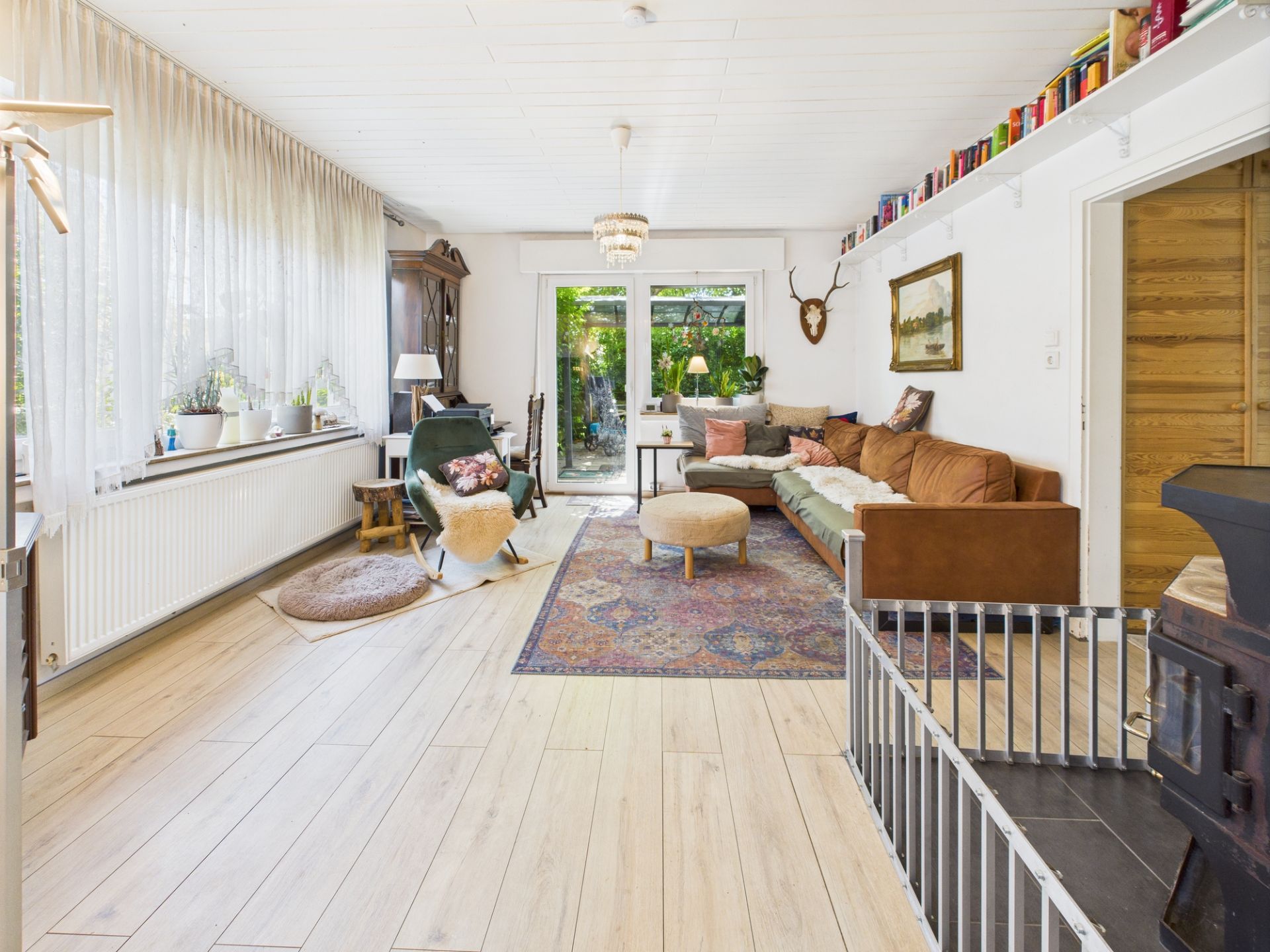Helles Wohnzimmer mit braunem Sofa, Sessel, Teppich und Blick auf den Garten durch Glastür.