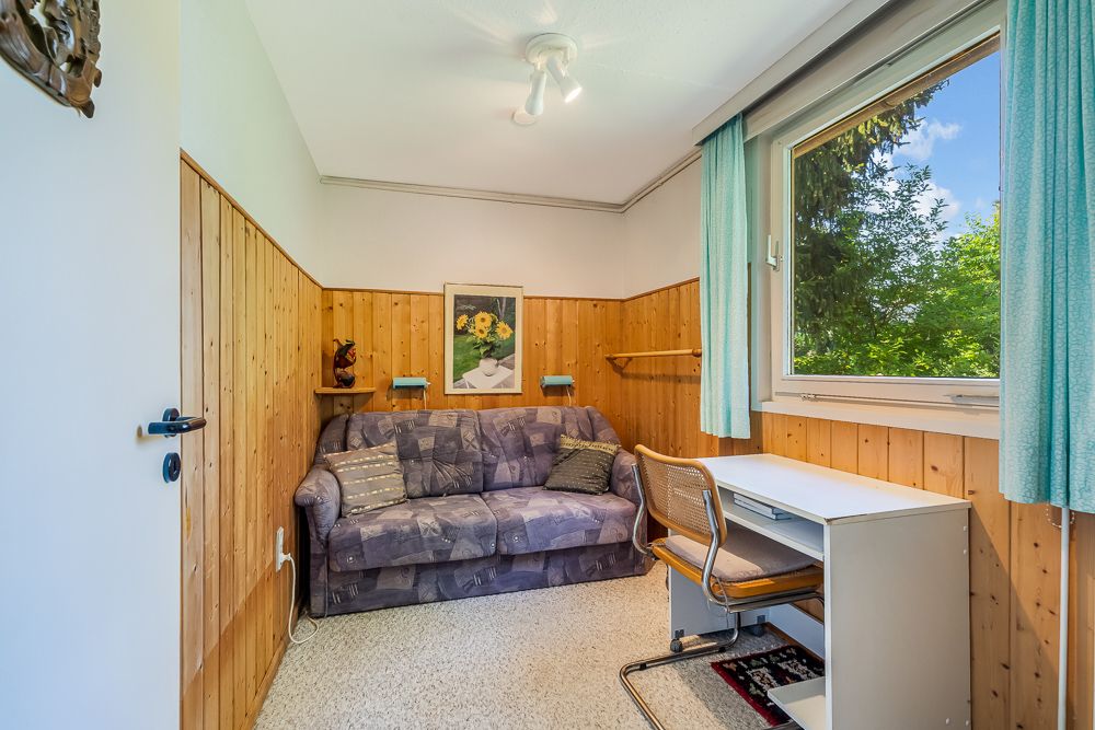 Kleines Zimmer mit Sofa, Schreibtisch, Stuhl und großem Fenster mit Blick ins Grüne.