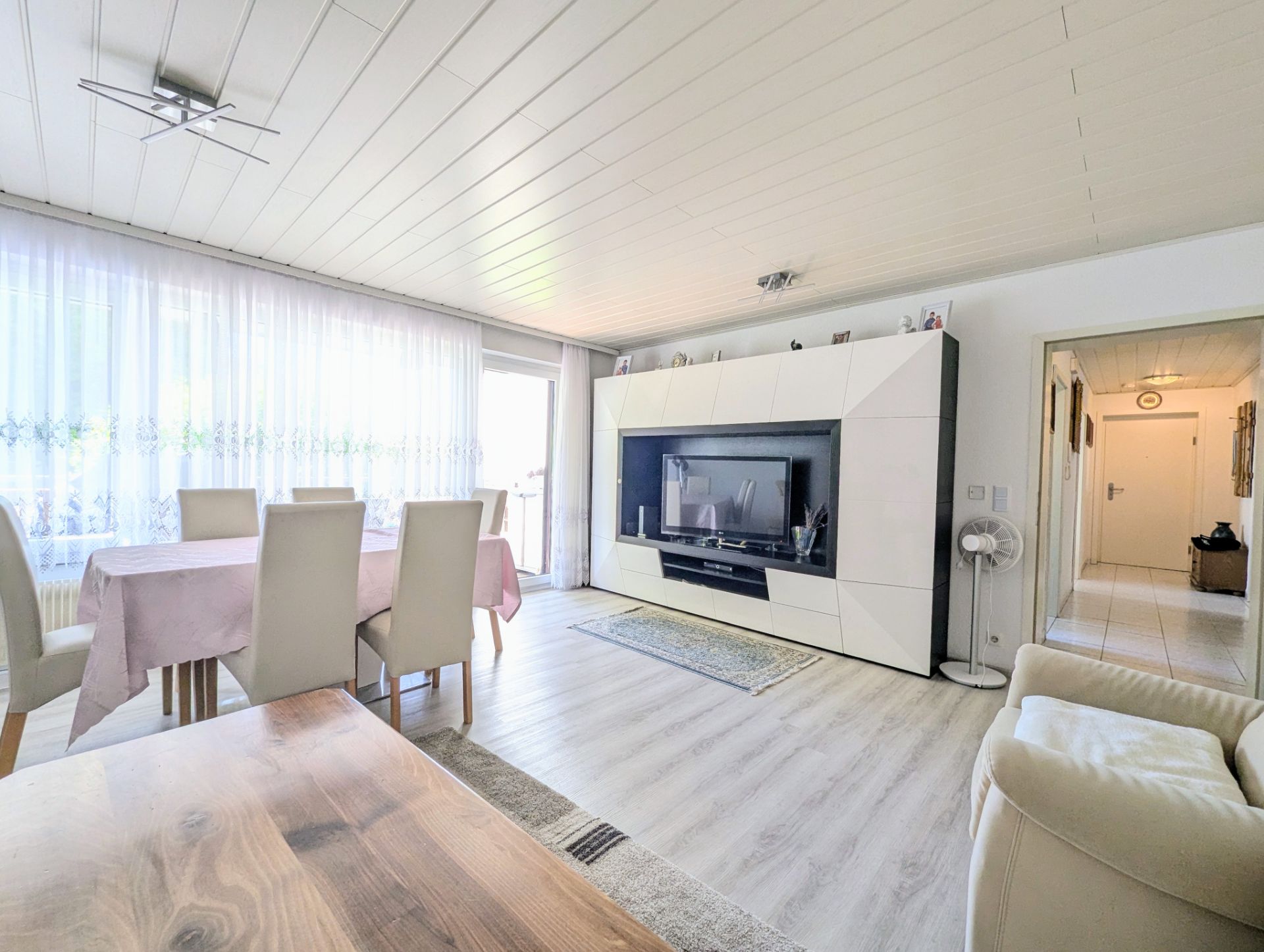 Helles Wohnzimmer mit Esstisch, Stühlen, TV-Wand und Sofa, großer Fensterfront mit Vorhängen.