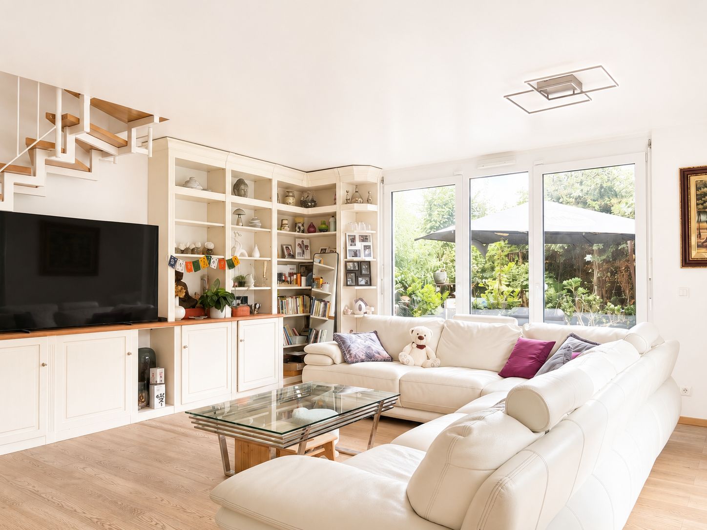 Helles Wohnzimmer mit weißem Sofa, Glastisch, großem Fernseher und Regalen vor großen Fenstern.