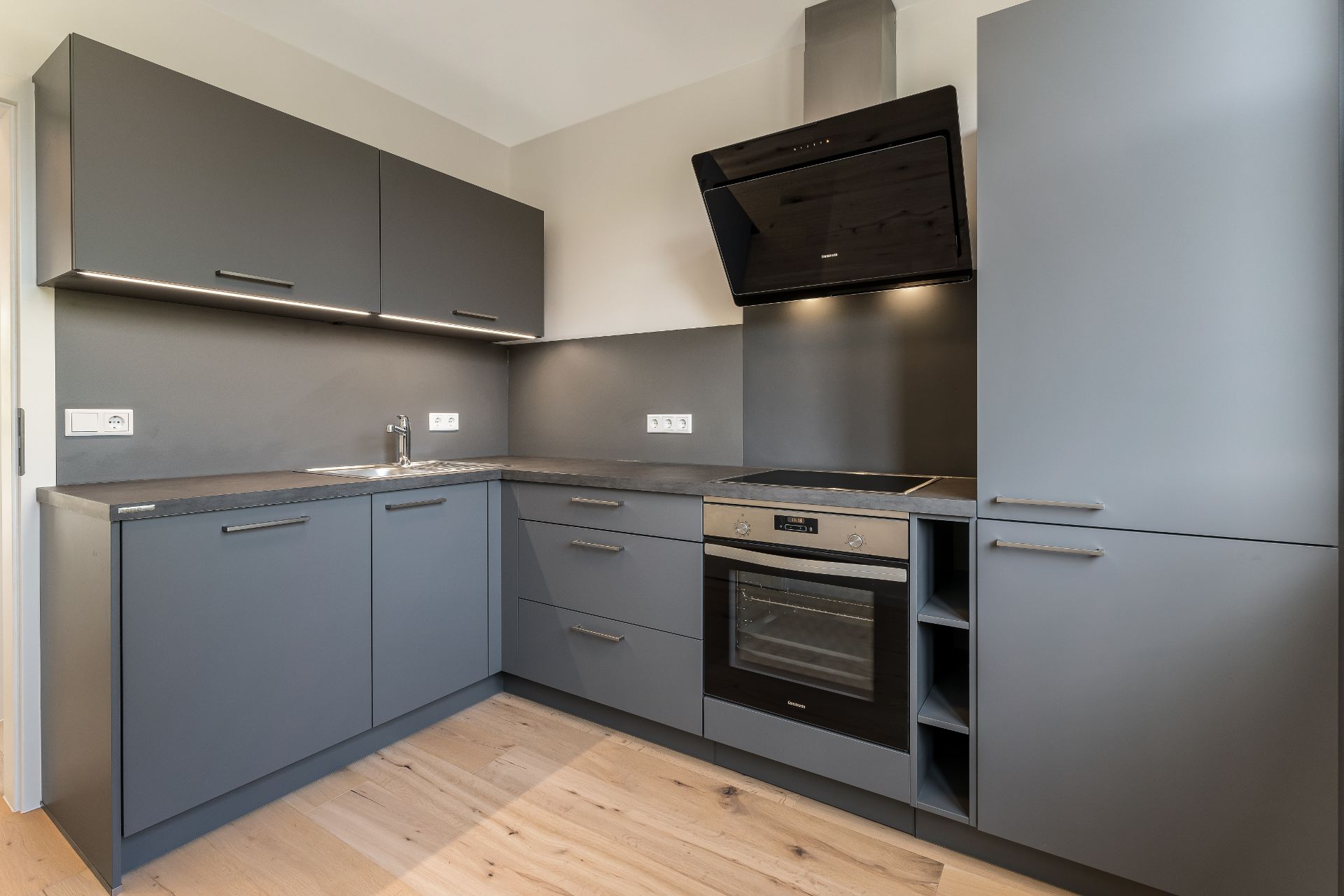 Moderne Einbauküche mit grauen Schränken, Backofen, Herd, Spüle und Holzfußboden.