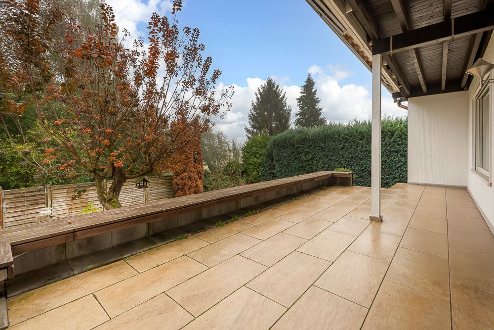 Überdachte Terrasse mit Fliesenboden, langer Bank, Baum und Hecke im Garten.