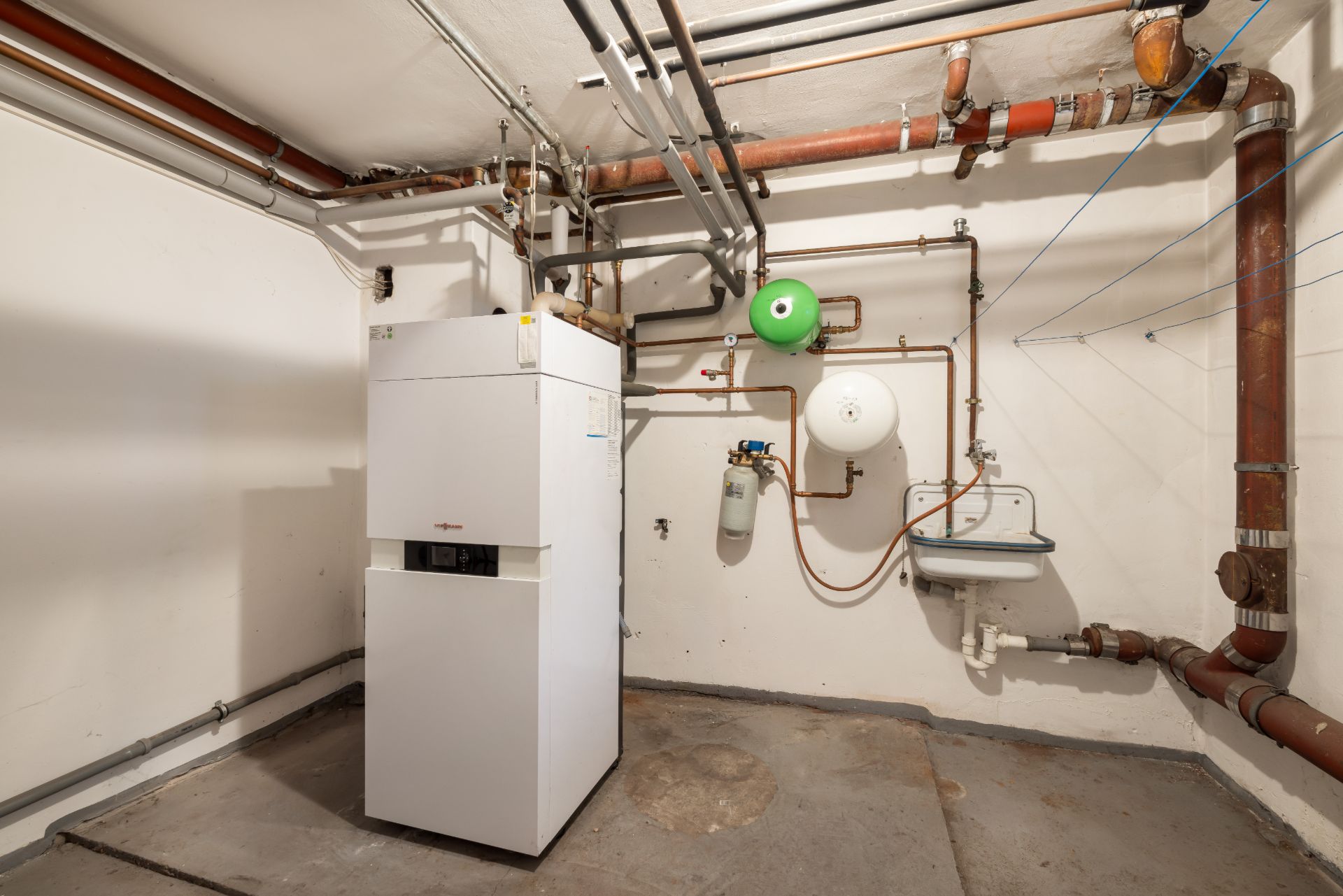 Heizungsraum mit Heizkessel, Rohren und kleinem Waschbecken an der Wand.