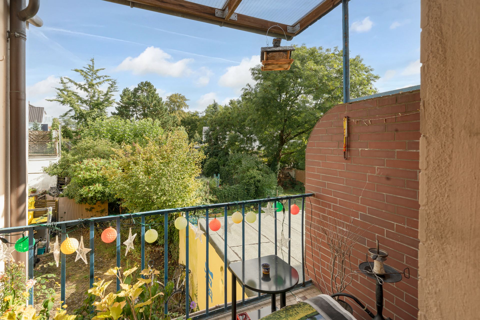 Balkon mit Lichterkette, kleinem Tisch und Pflanzen, Blick auf grünen Garten.