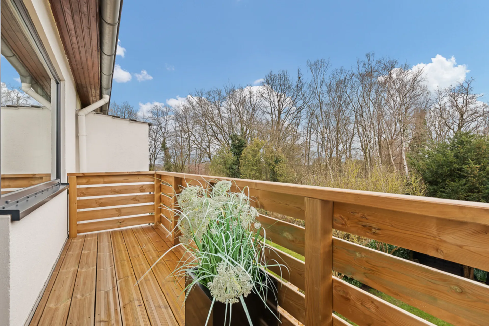 Holzbalkon mit Blumenkasten und Blick auf Bäume im Hintergrund bei blauem Himmel.