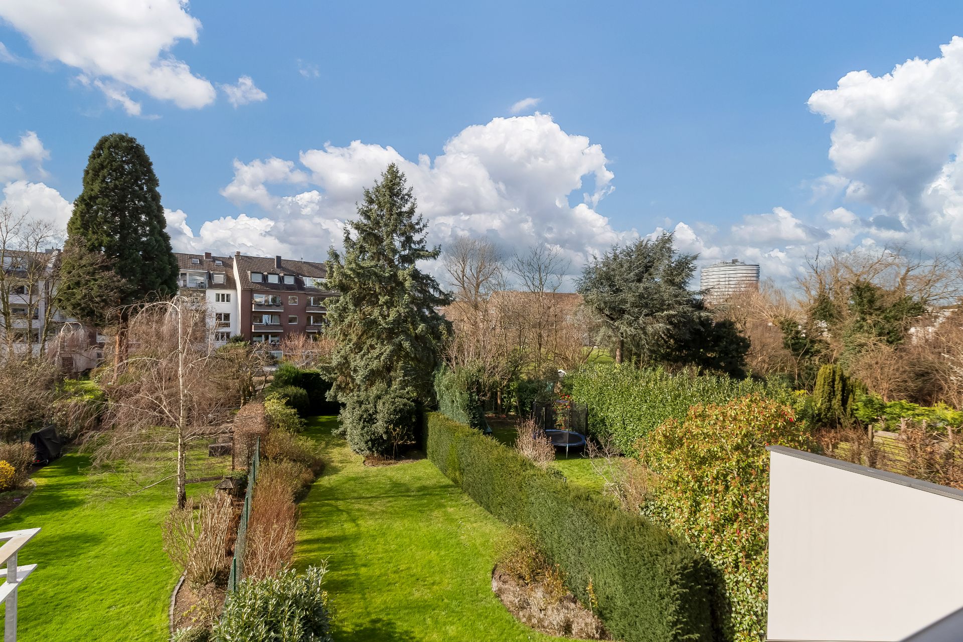 Grüner Garten mit Bäumen und Hecken, im Hintergrund Wohnhäuser unter blauem Himmel.