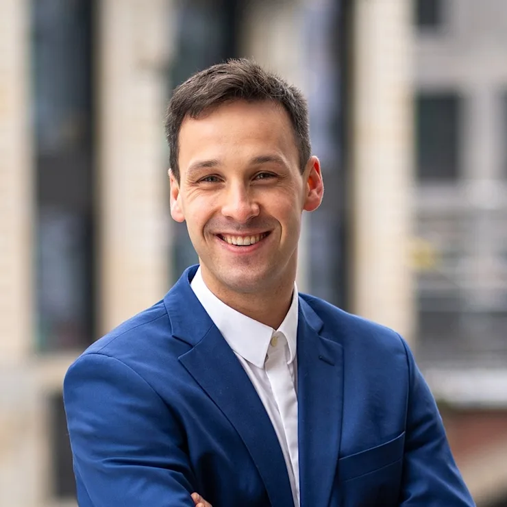 Ein Mann in blauem Anzug steht draußen mit verschränkten Armen und lächelt in die Kamera.