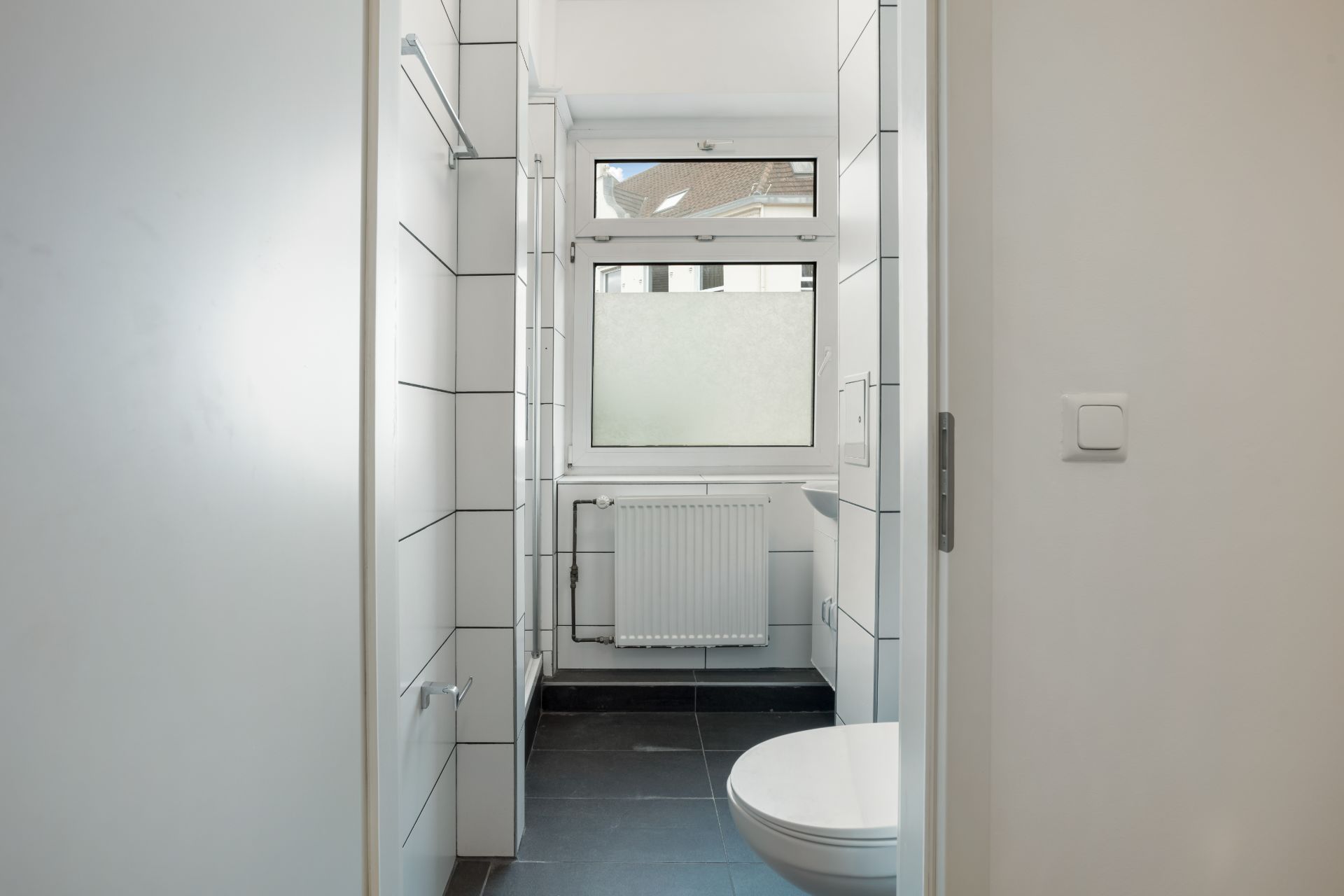 Helles Badezimmer mit weiß gefliesten Wänden, Fenster, Heizung und Toilette.