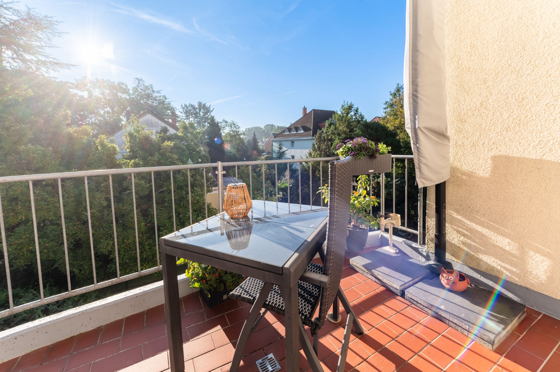Kleiner Balkon mit Glastisch, zwei Stühlen und Pflanzen, Blick auf Bäume und Nachbarhäuser.