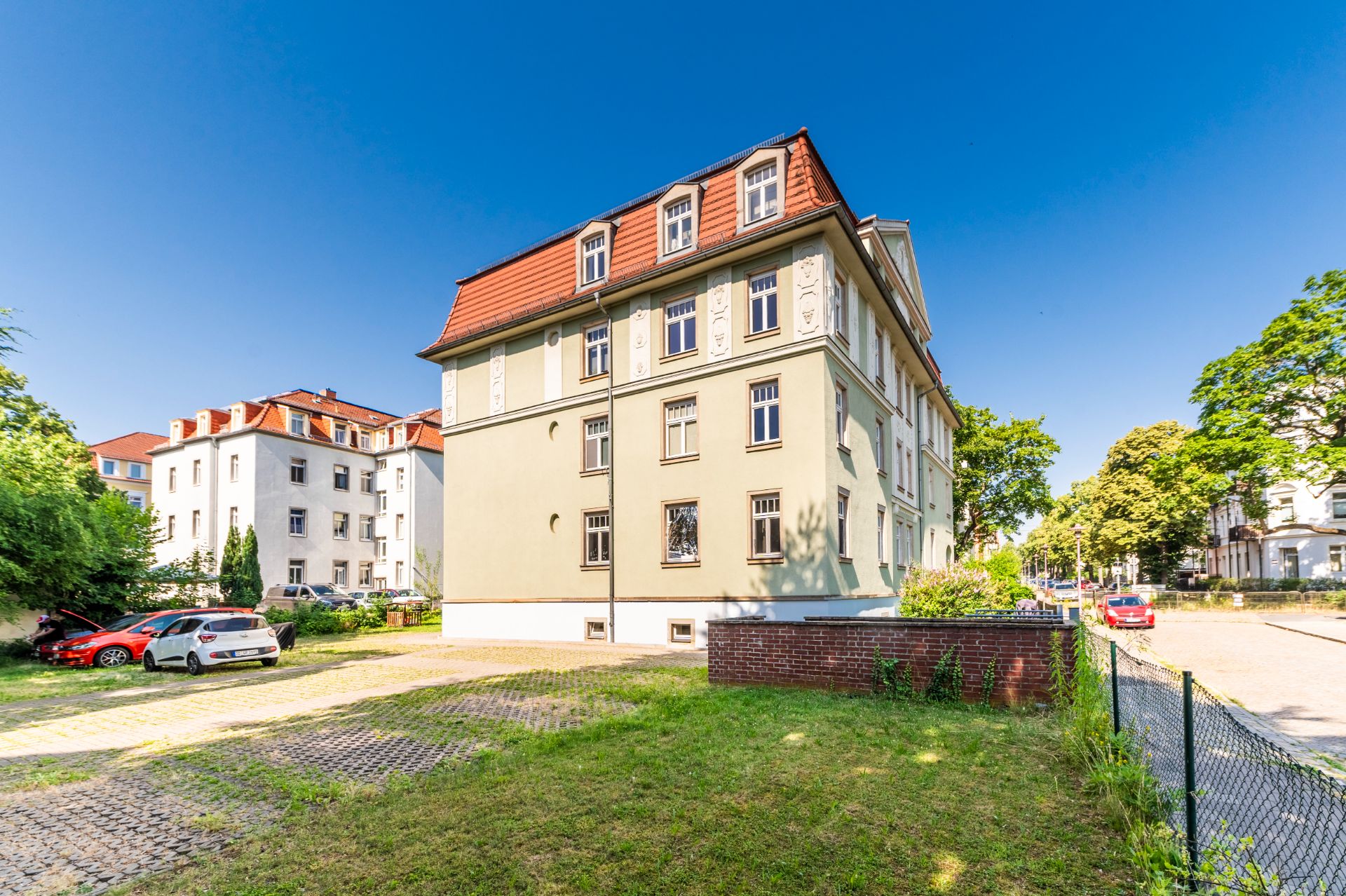 Mehrfamilienhaus mit rotem Dach, Grünfläche und Parkplatz im Vordergrund, bei sonnigem Wetter.