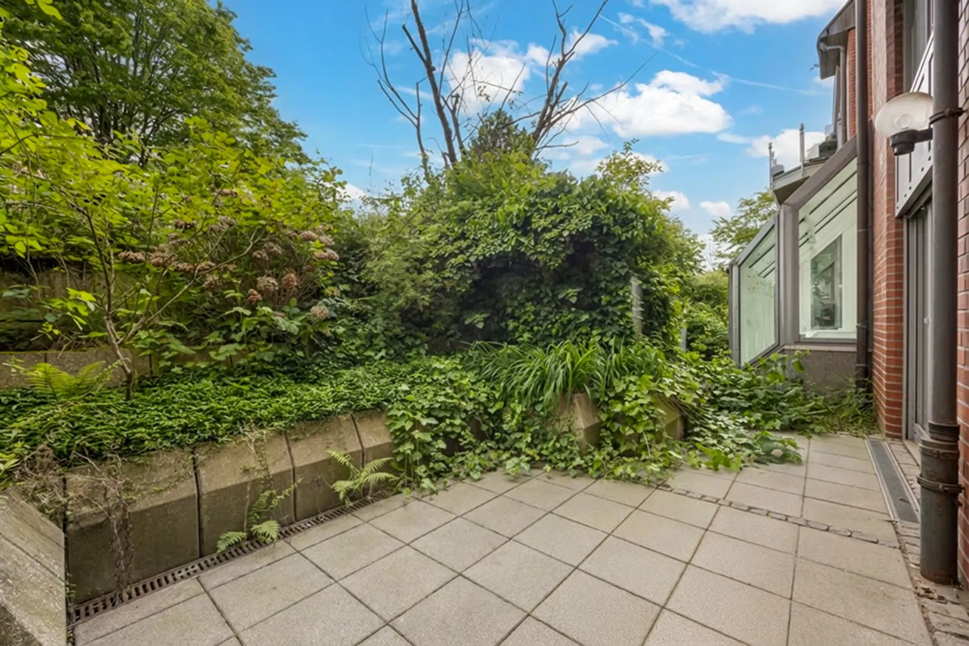 Gepflasterte Terrasse mit niedriger Mauer und viel Grünbewuchs, angrenzend an ein Wohnhaus.