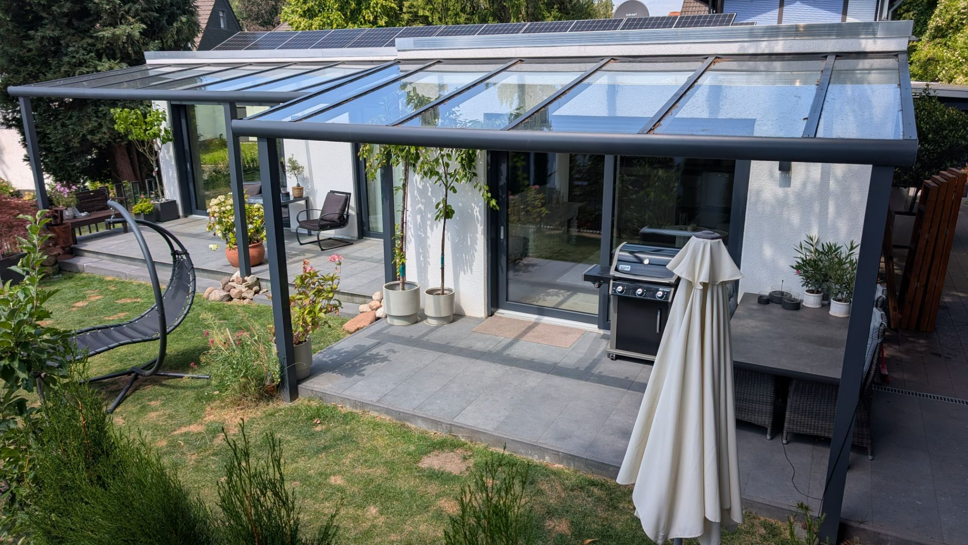 Moderne Terrasse mit Glasdach, Grill, Pflanzen und Gartenmöbeln an einem Wohnhaus.
