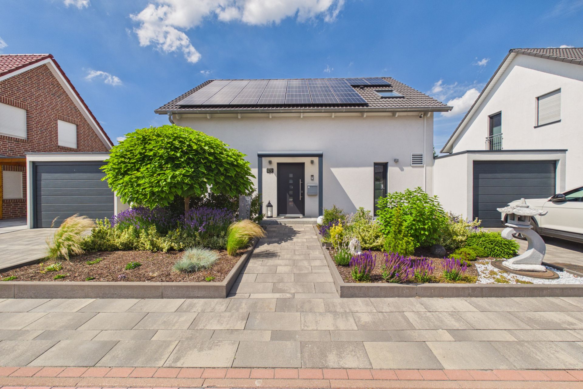 Modernes Einfamilienhaus mit Solardach, Vorgarten und zwei Garagen, unter blauem Himmel.