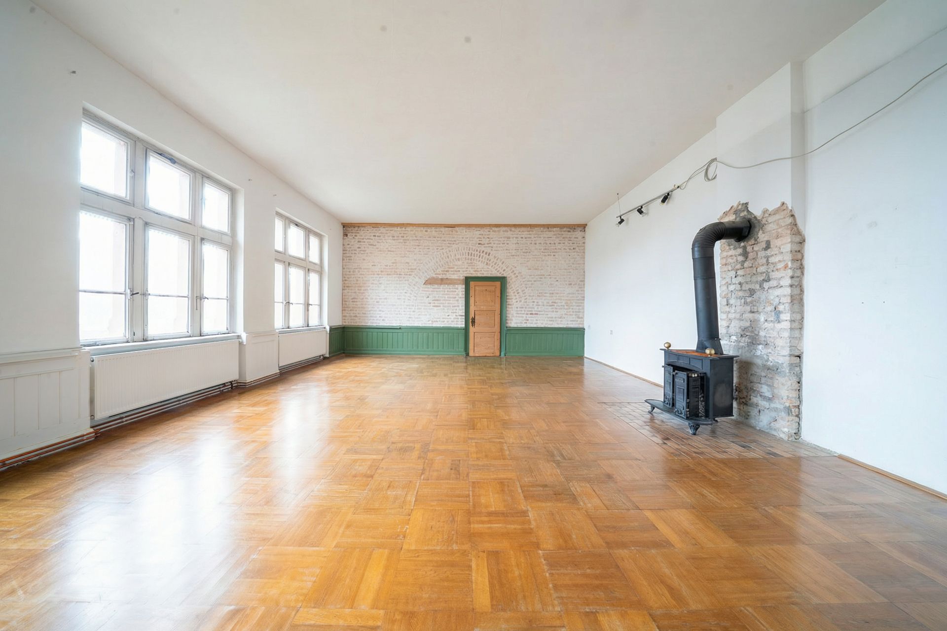 Heller Raum mit Holzfußboden, großen Fenstern und einem Kaminofen an der rechten Wand.