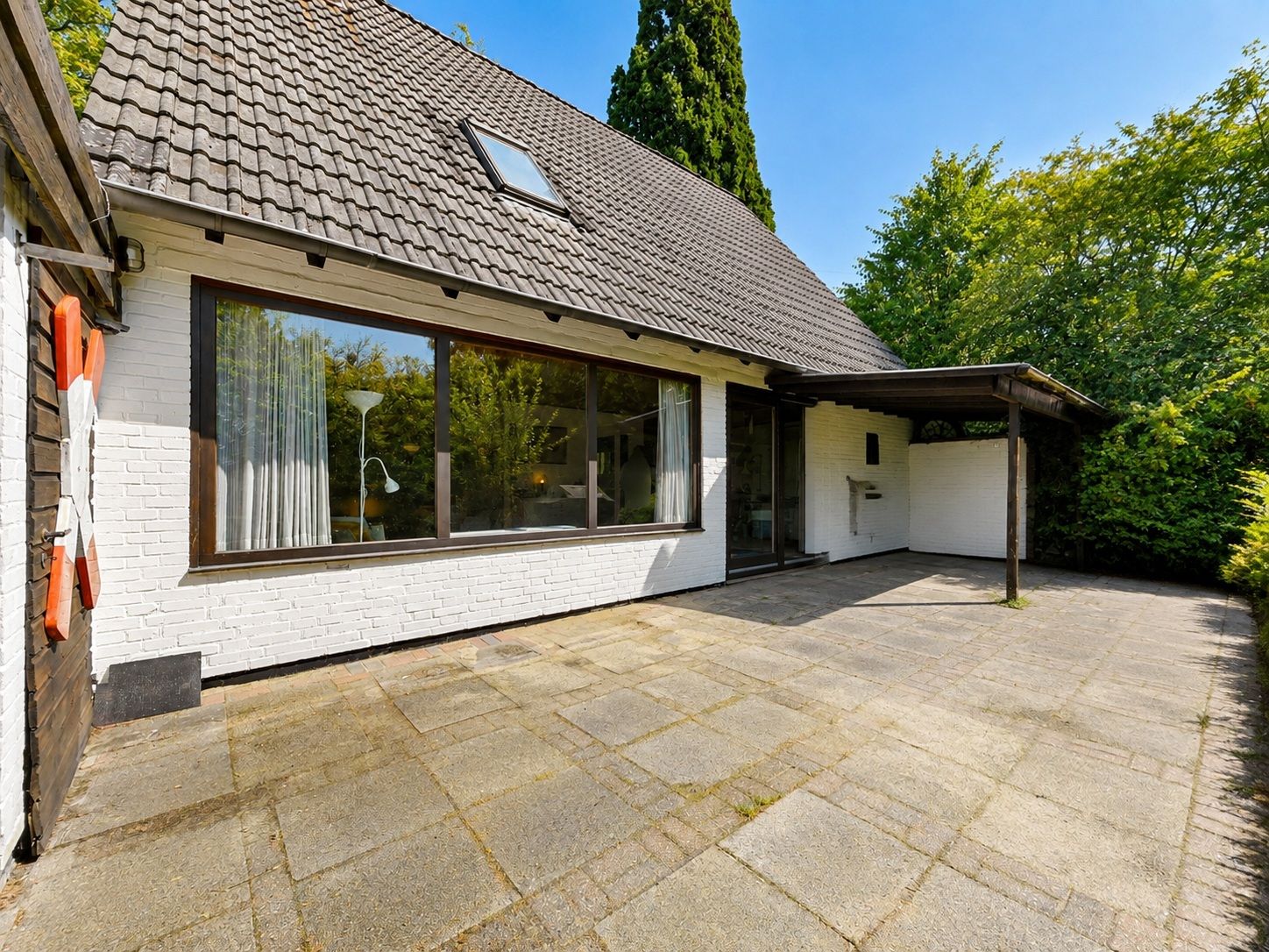 Große gepflasterte Terrasse vor einem weißen Wohnhaus mit großem Fenster und schrägem Dach.