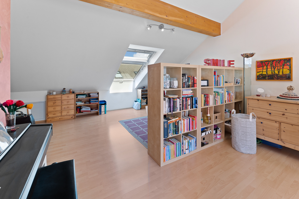 Helles Dachzimmer mit Bücherregal, Holzkommoden, Klavier und Dachfenster.