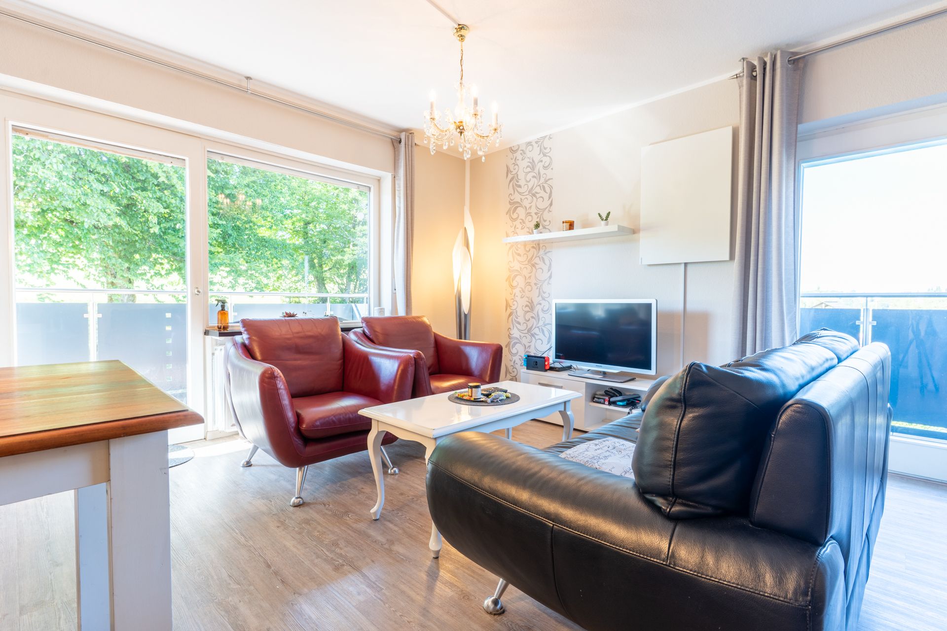 Helles Wohnzimmer mit zwei roten Sesseln, schwarzem Sofa, Couchtisch und Fernseher, große Fenster mit Blick ins Grüne.