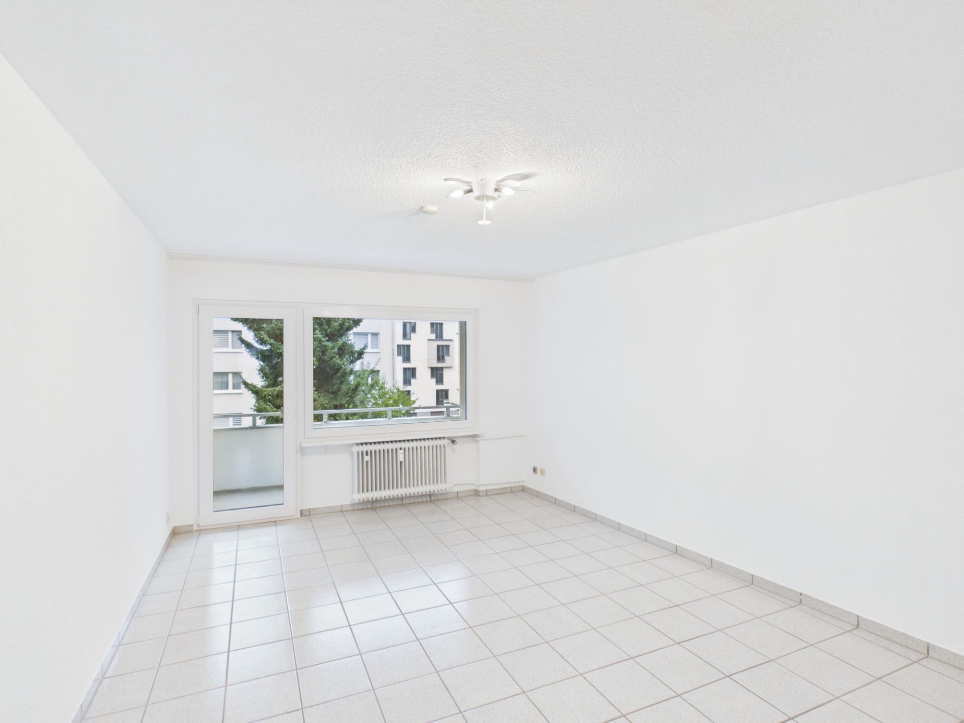 Helles, leeres Zimmer mit Fliesenboden, weißem Wänden und großem Fenster mit Blick nach draußen.