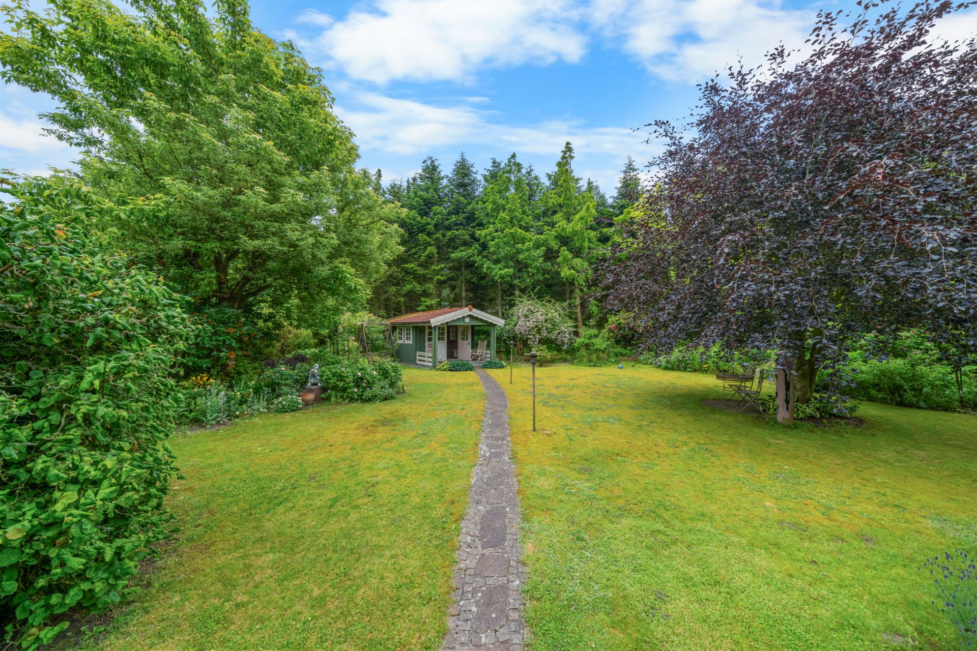 Gepflasterter Weg führt durch grünen Garten zu kleinem Haus mit Bäumen im Hintergrund.