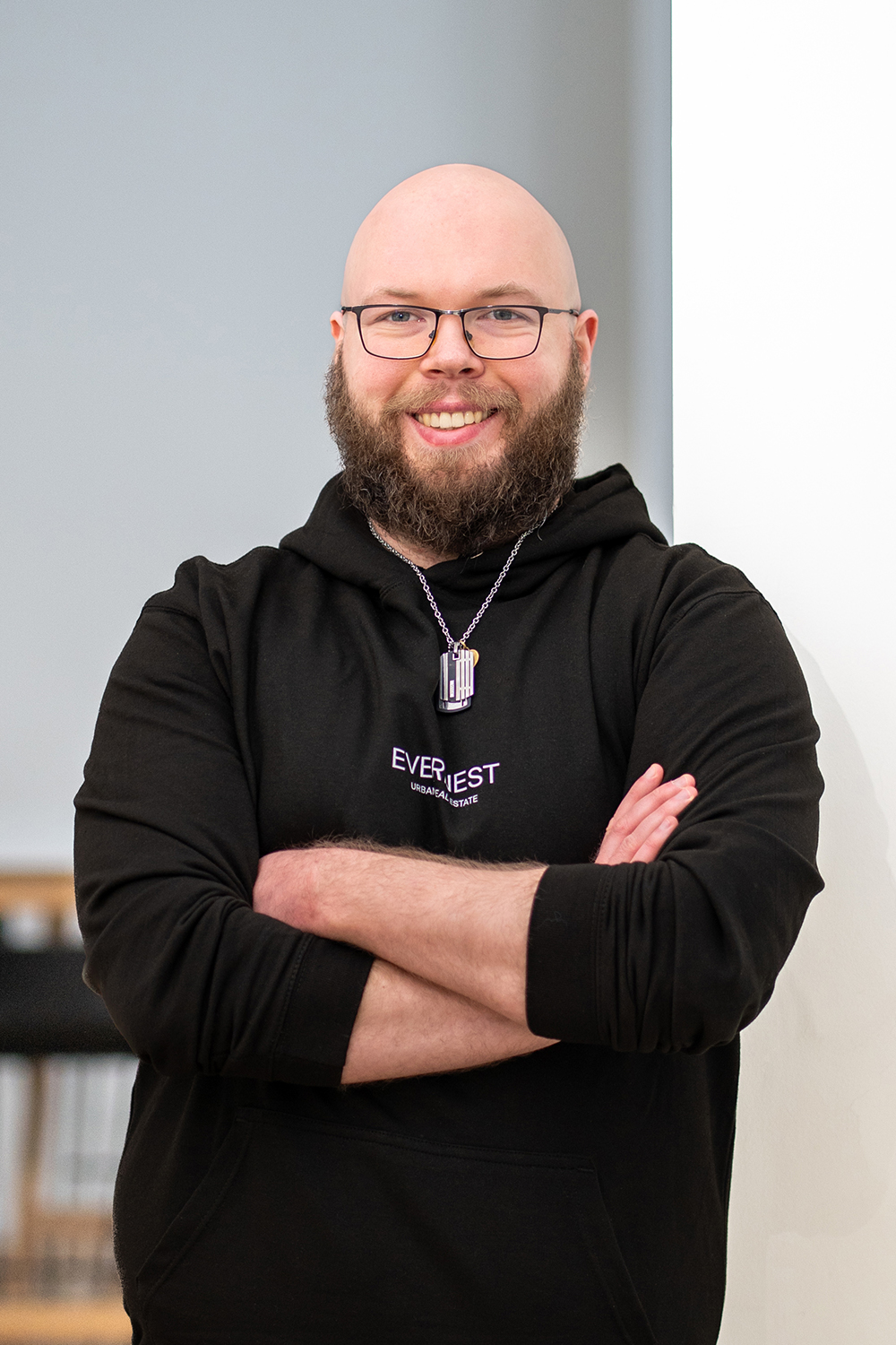 Ein bärtiger Mann mit Brille und schwarzem Hoodie steht mit verschränkten Armen in einem Raum.