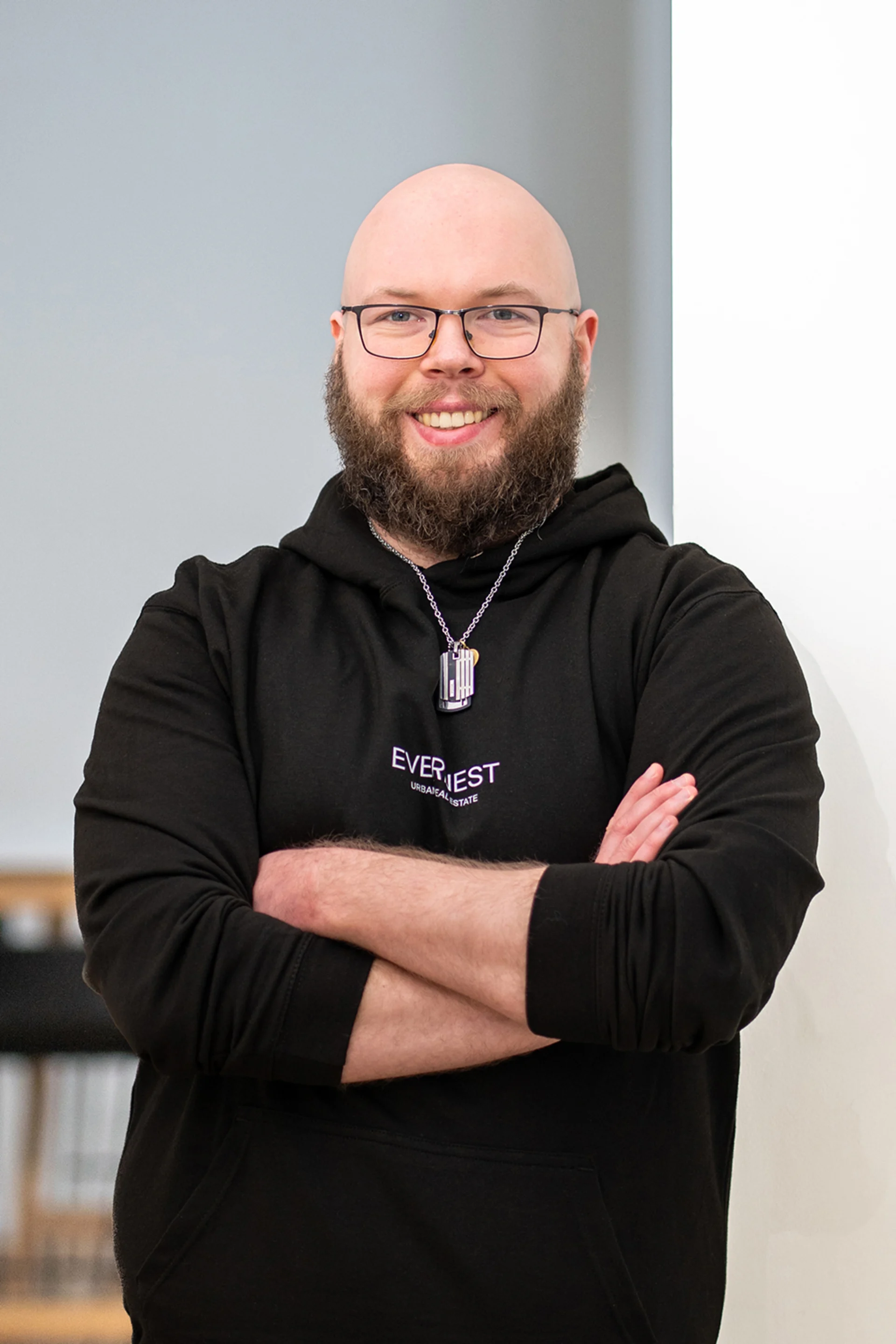 Ein bärtiger Mann mit Brille und schwarzem Hoodie steht mit verschränkten Armen in einem Raum.