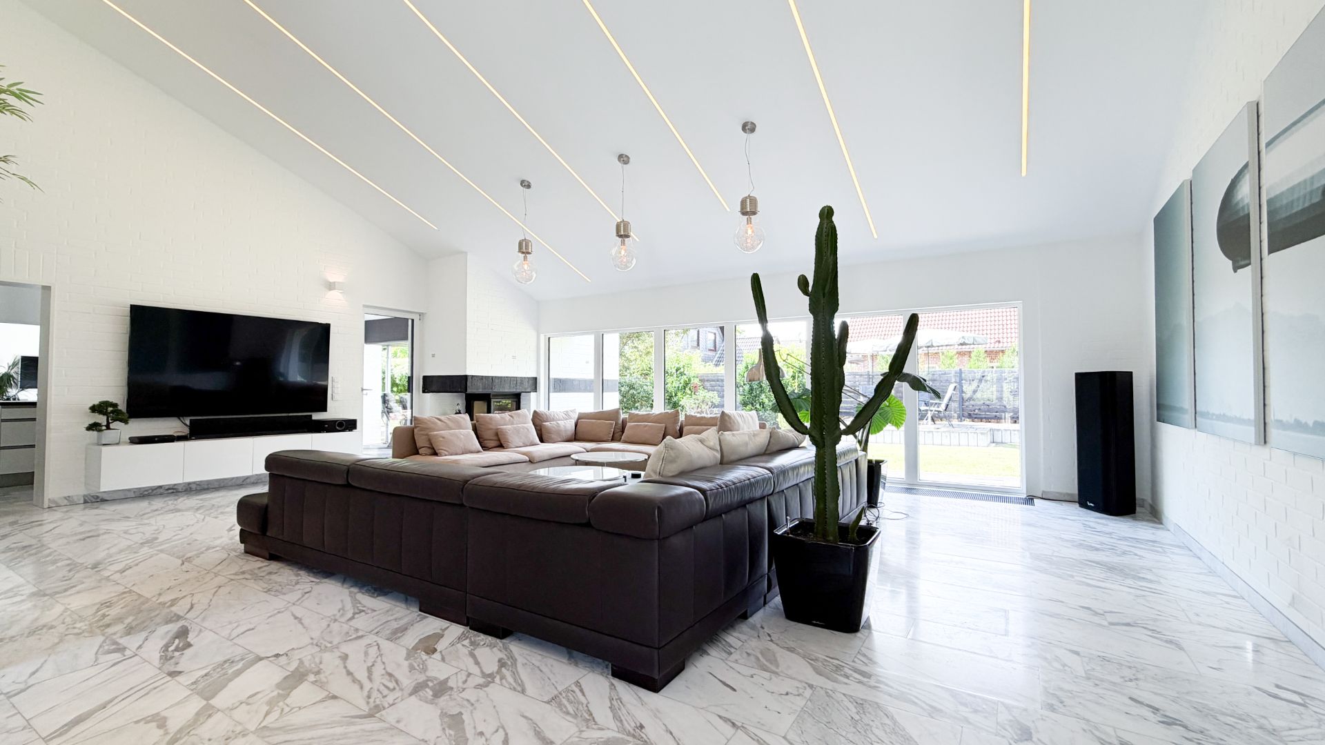 Modernes Wohnzimmer mit großer dunkler Eckcouch, Marmorboden, Fernseher und einem großen Kaktus.