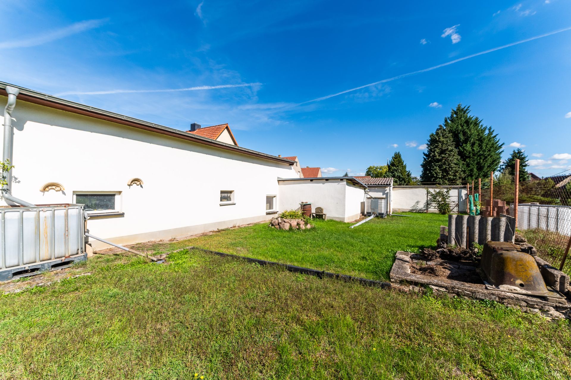 Grüner Garten mit Rasen neben einem weißen Haus, einige Bäume und Beete im Hintergrund.