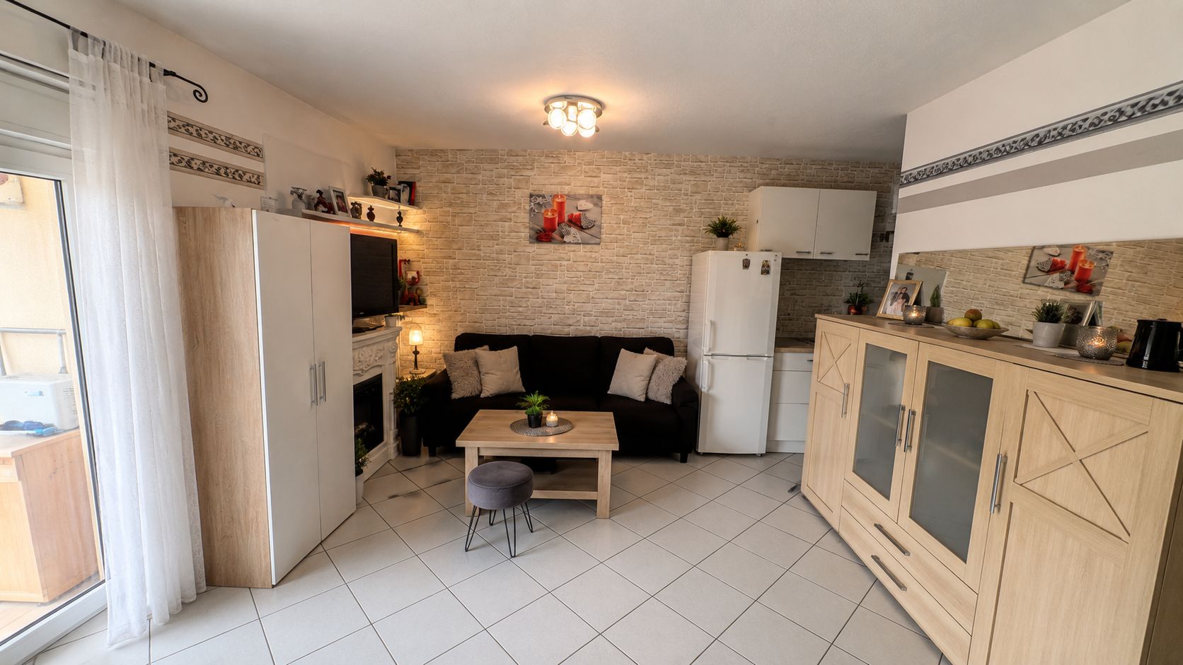 Wohnzimmer mit schwarzem Sofa, Holzmöbeln, Kühlschrank und kleinem Couchtisch auf Fliesenboden.