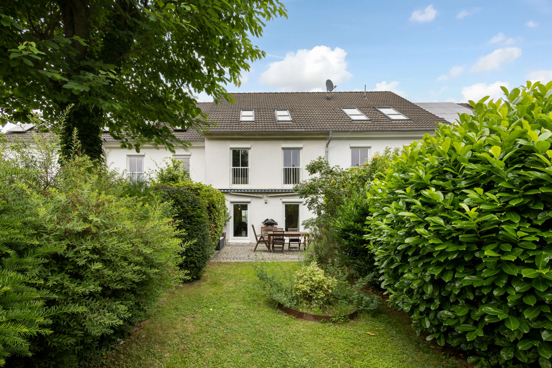 Garten mit Rasen, Terrasse, Gartenmöbeln und Blick auf ein weißes Reihenhaus.