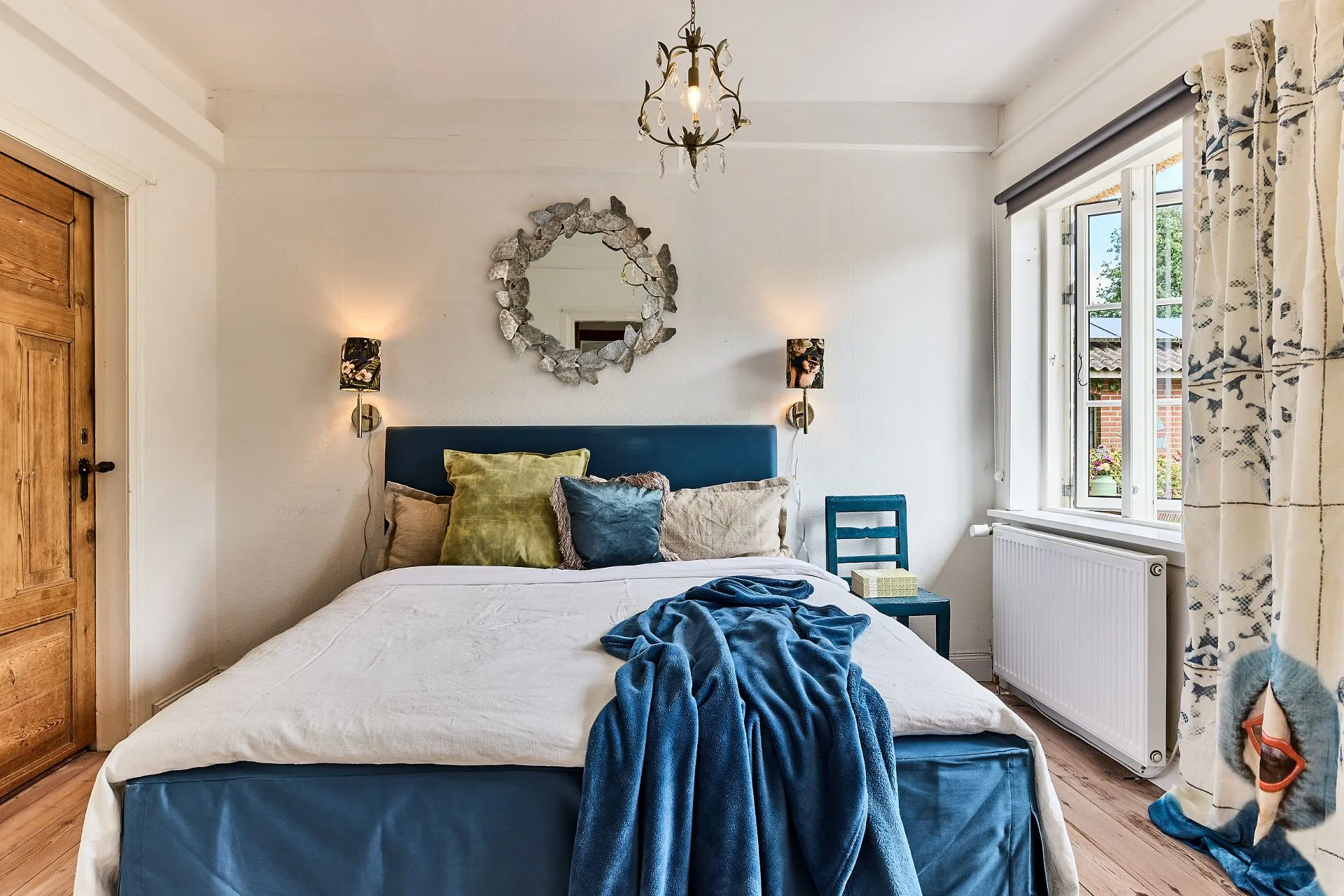 Helles Schlafzimmer mit Doppelbett, bunten Kissen, Spiegel, Holzstuhl und großem Fenster.