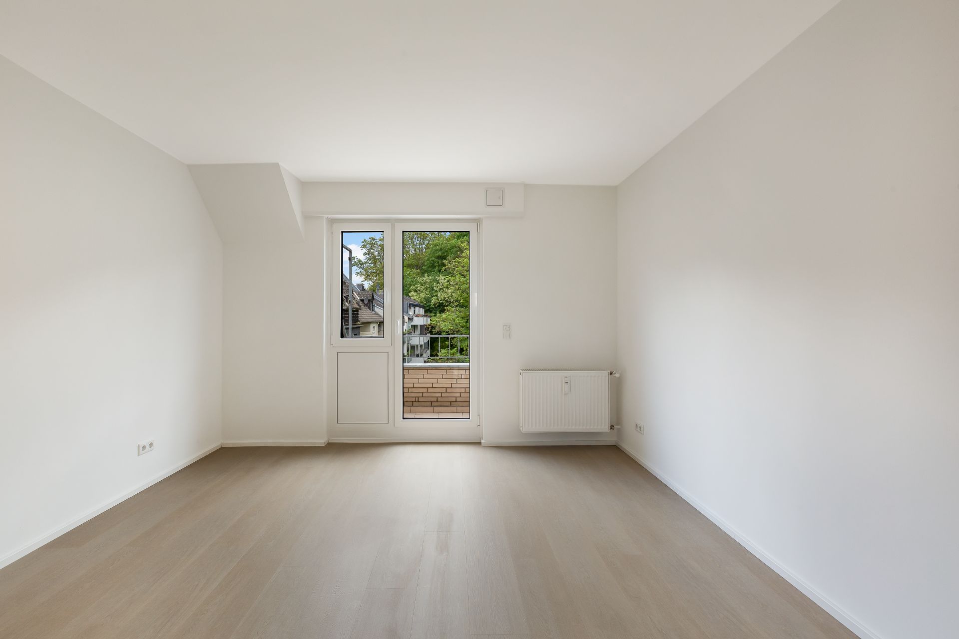 Helles, leeres Zimmer mit Holzboden, weißen Wänden und Tür zum Balkon.