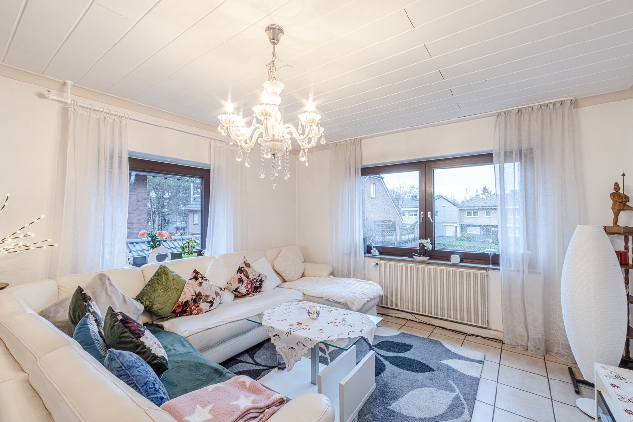 Helles Wohnzimmer mit weißem Ecksofa, vielen Kissen, Kronleuchter und zwei großen Fenstern.