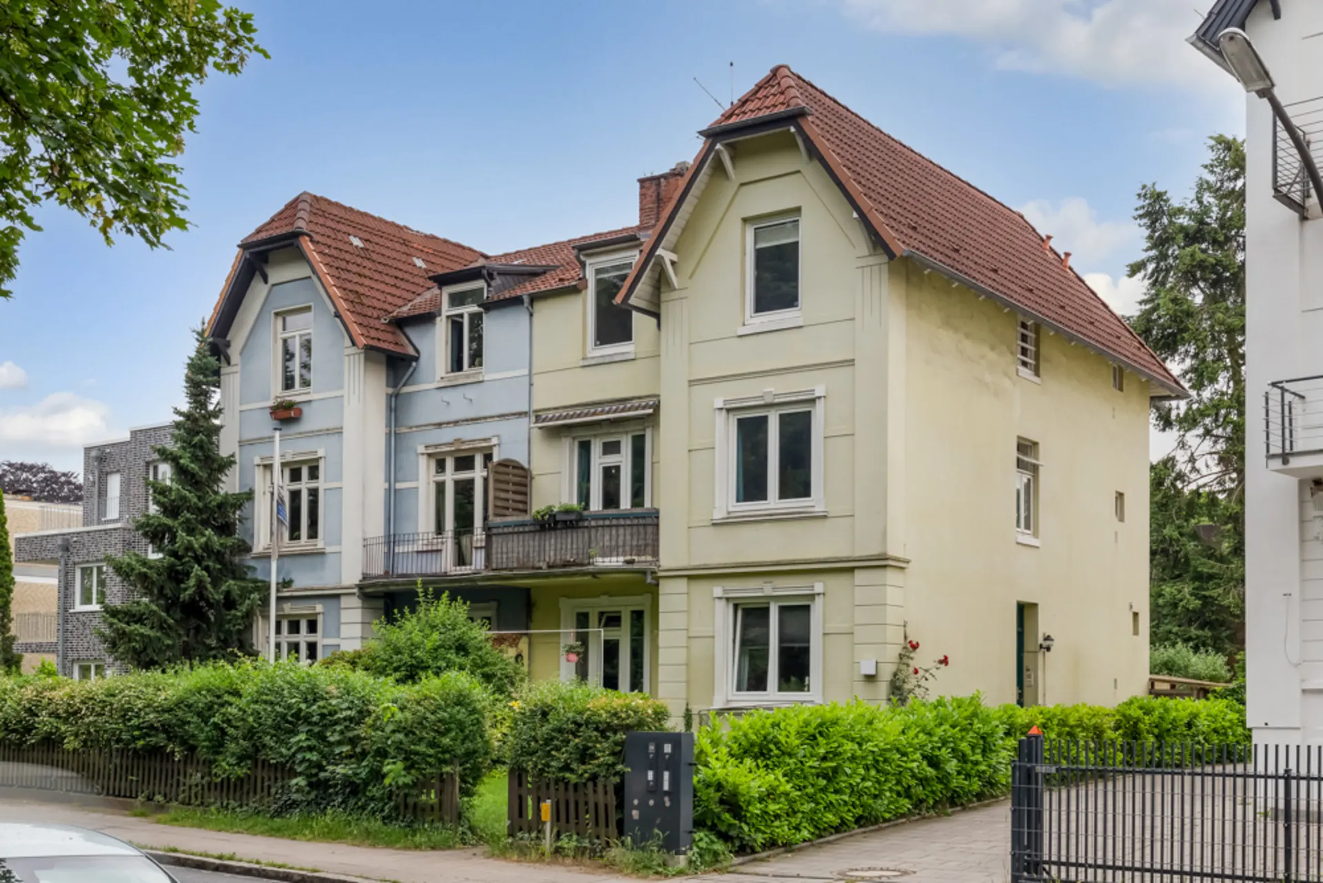Dreistöckiges Wohnhaus mit rotem Dach und kleinem Garten, umgeben von Büschen und Zaun.