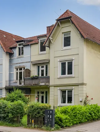 Dreistöckiges Wohnhaus mit rotem Dach und kleinem Garten, umgeben von Büschen und Zaun.