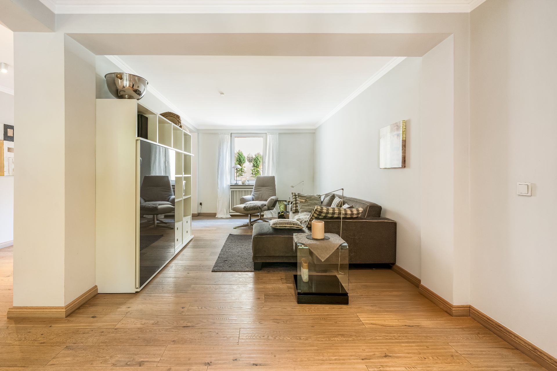 Helles Wohnzimmer mit Holzfußboden, Sofa, Sessel, Regal und Fenster mit weißen Vorhängen.
