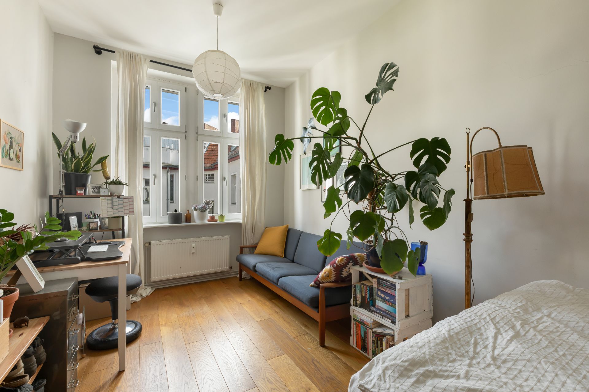 Helles Zimmer mit Holzfußboden, Schreibtisch, blauem Sofa, großem Fenster und vielen Pflanzen.