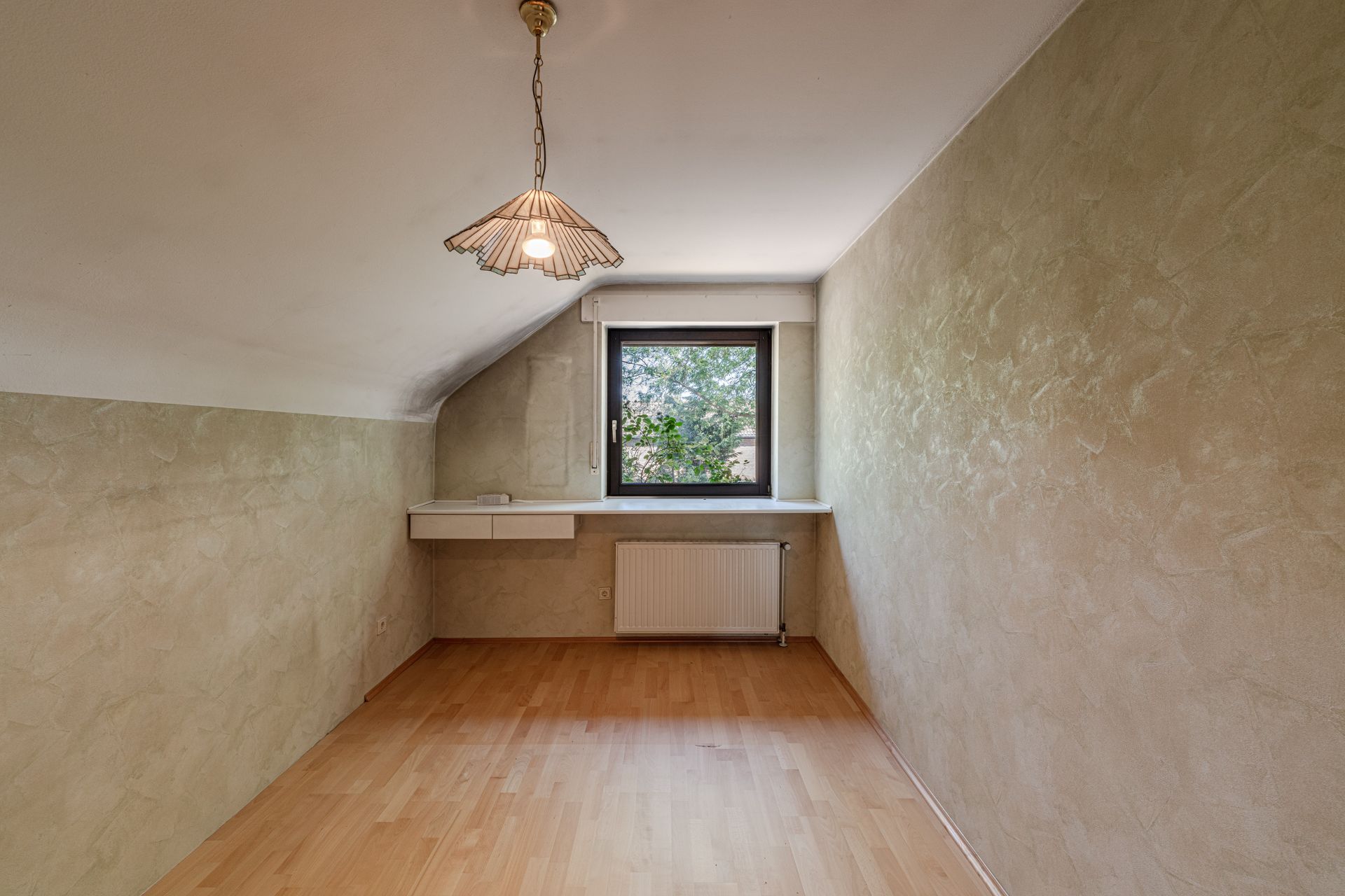 Kleines Zimmer mit Holzfußboden, Dachschräge, einem Fenster und einer Deckenlampe.