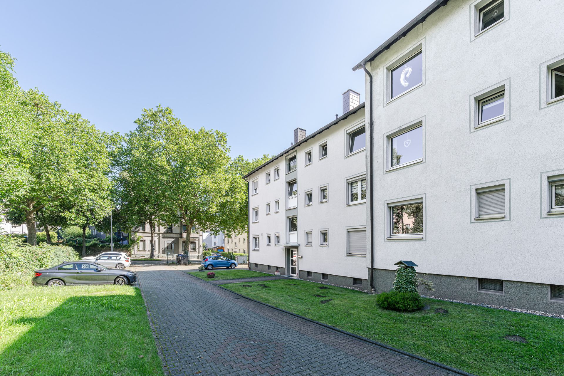 Mehrfamilienhaus mit weißer Fassade, Grünfläche und parkenden Autos bei Tageslicht.