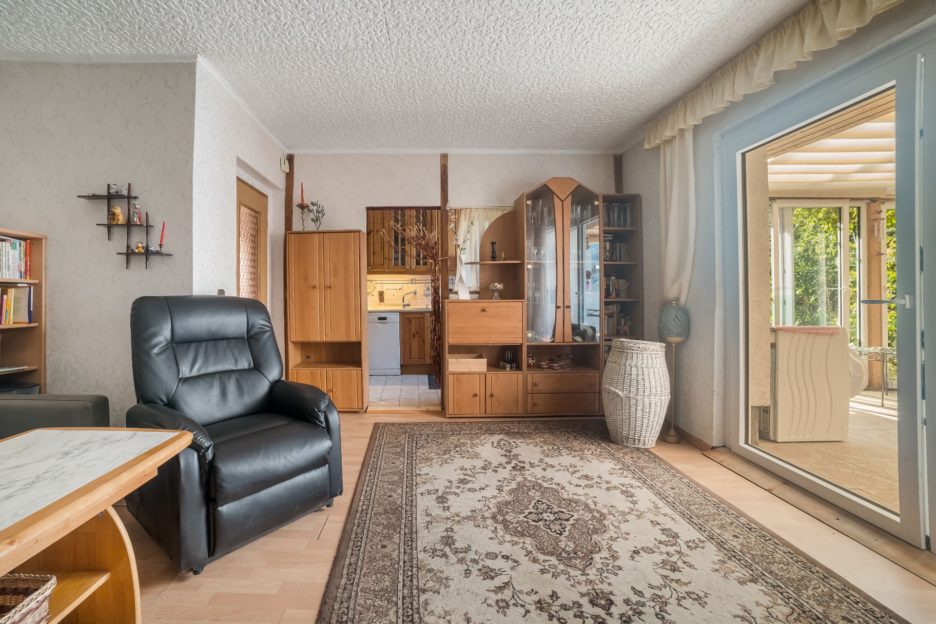 Wohnzimmer mit schwarzem Sessel, Holzmöbeln, Teppich und Zugang zu einem hellen Wintergarten.