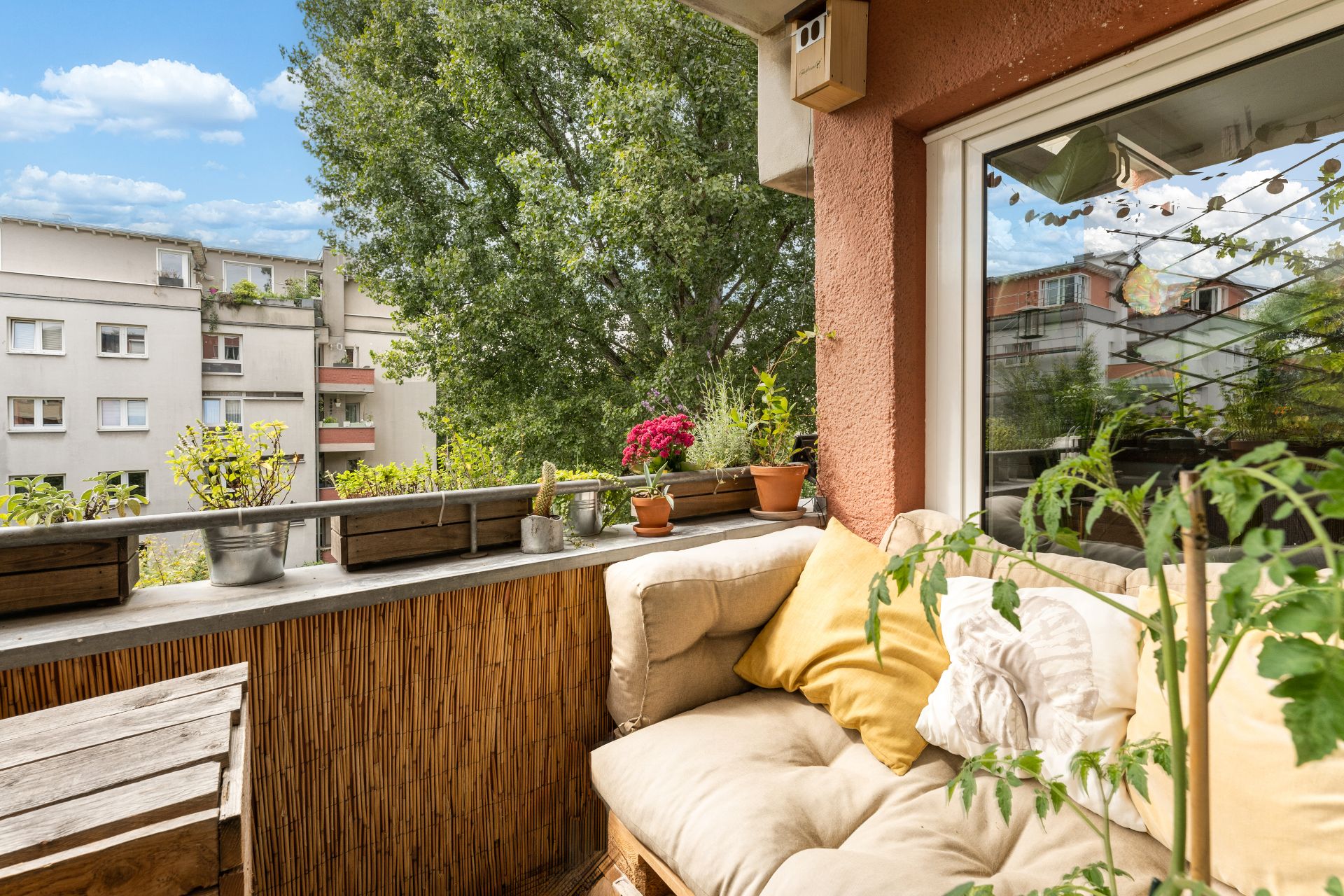 Gemütlicher Balkon mit Sitzbank, gelben Kissen, Pflanzen und Blick auf Bäume und Nachbargebäude.