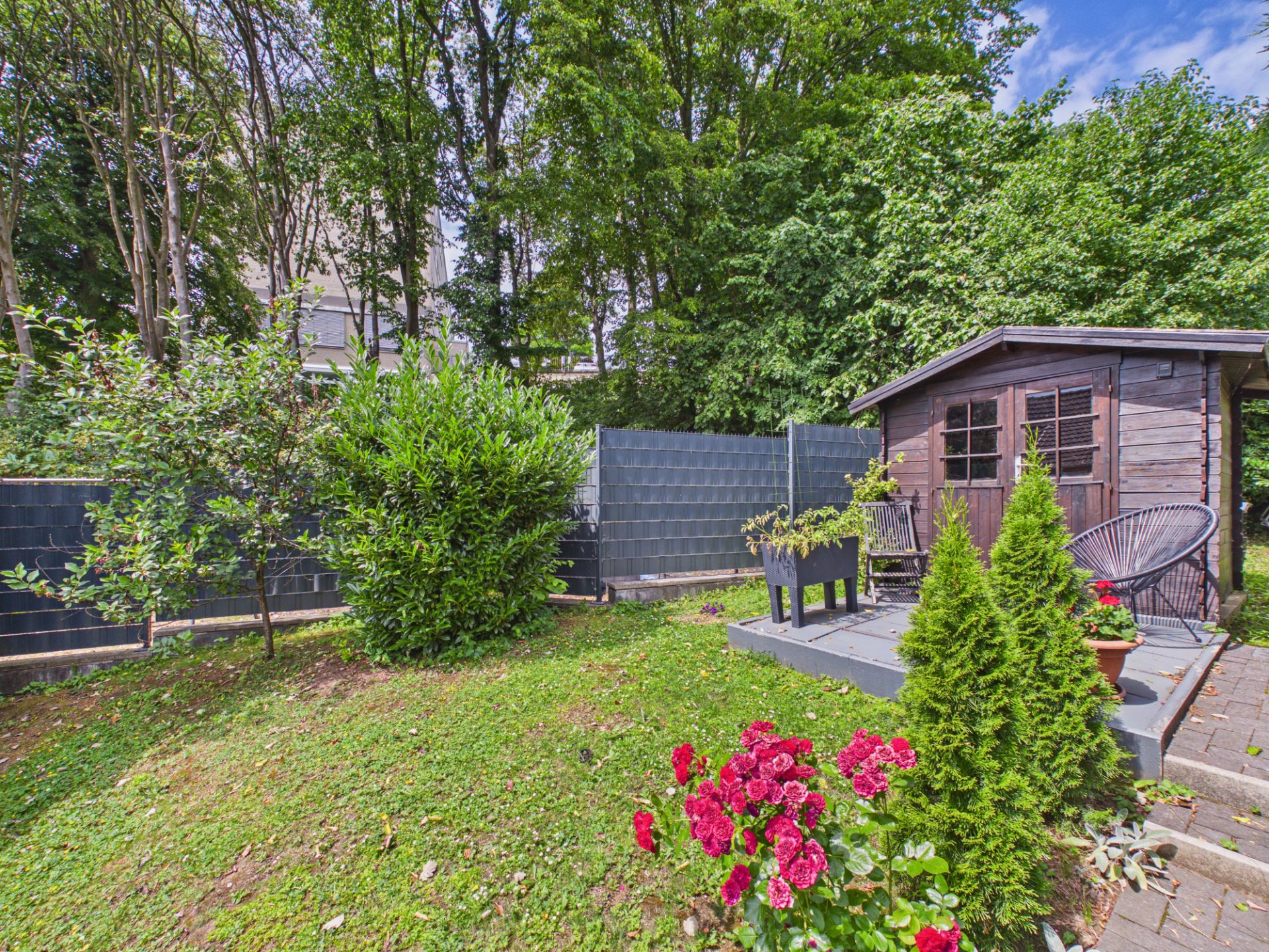 Garten mit bunten Blumen, Sträuchern und einem kleinen Holzhaus auf einer Terrasse.