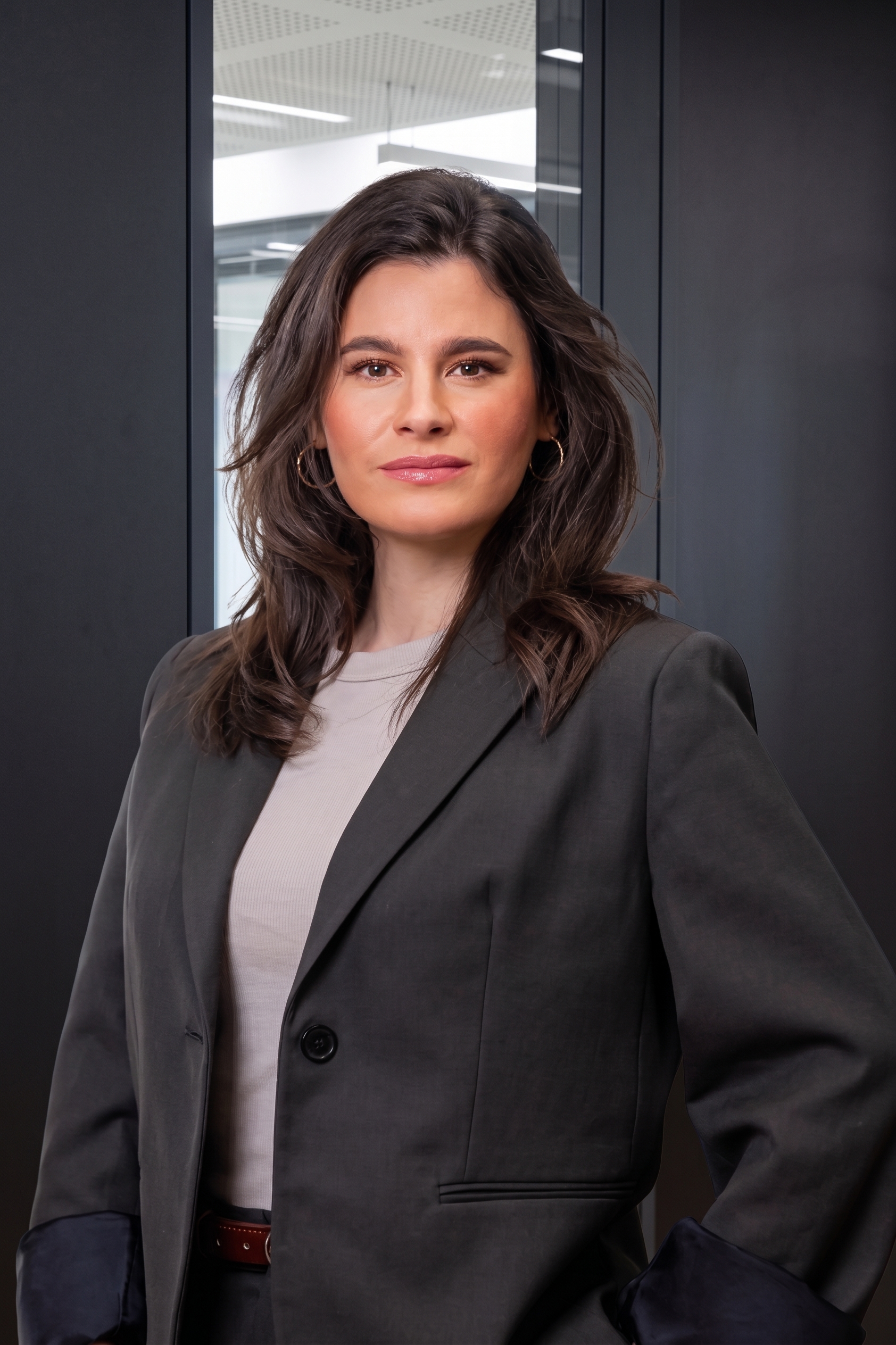 Frau mit braunen Haaren im Blazer steht vor einer Glastür in einem modernen Raum.