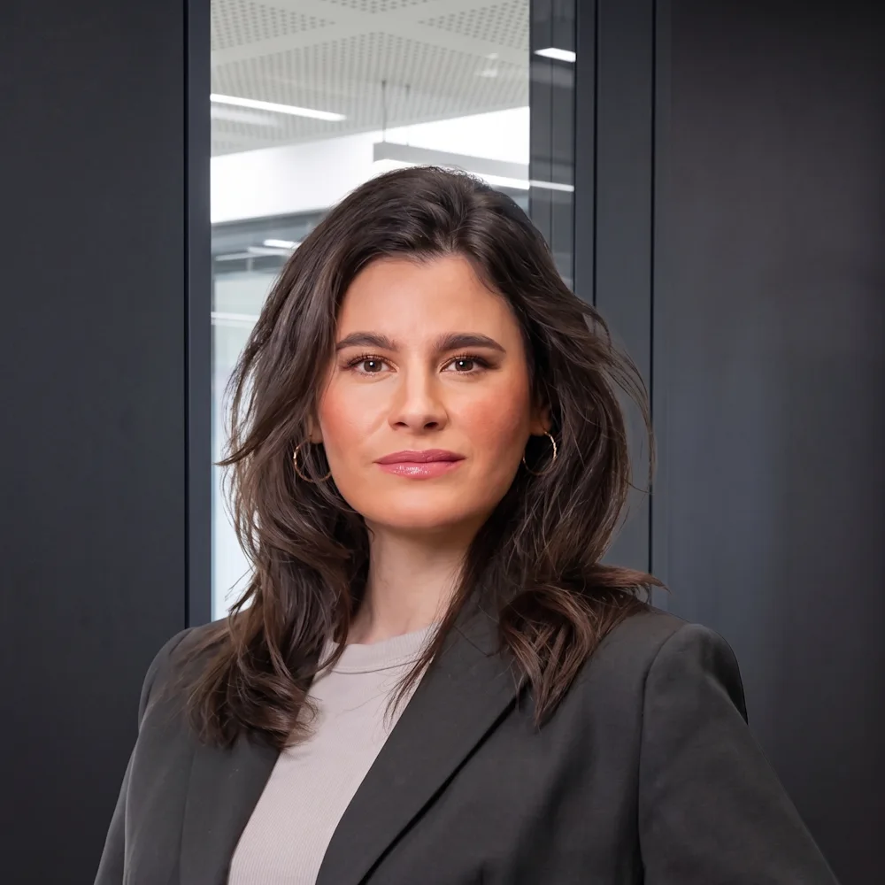 Frau mit braunen Haaren im Blazer steht vor einer Glastür in einem modernen Raum.