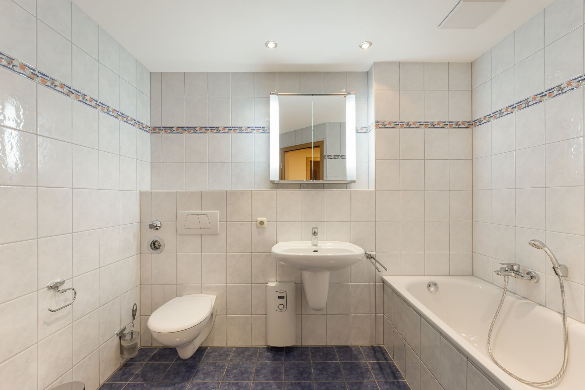 Modernes Badezimmer mit Badewanne, Wandtoilette, Waschbecken und Spiegel auf blau gefliestem Boden.