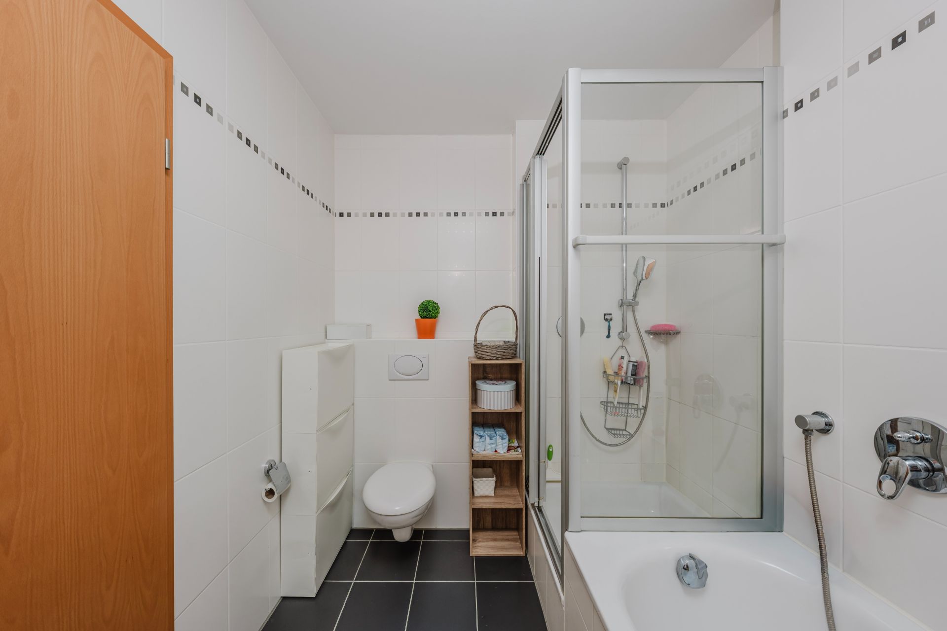 Modernes Badezimmer mit Badewanne, Dusche, WC und Regal auf dunklem Fliesenboden.